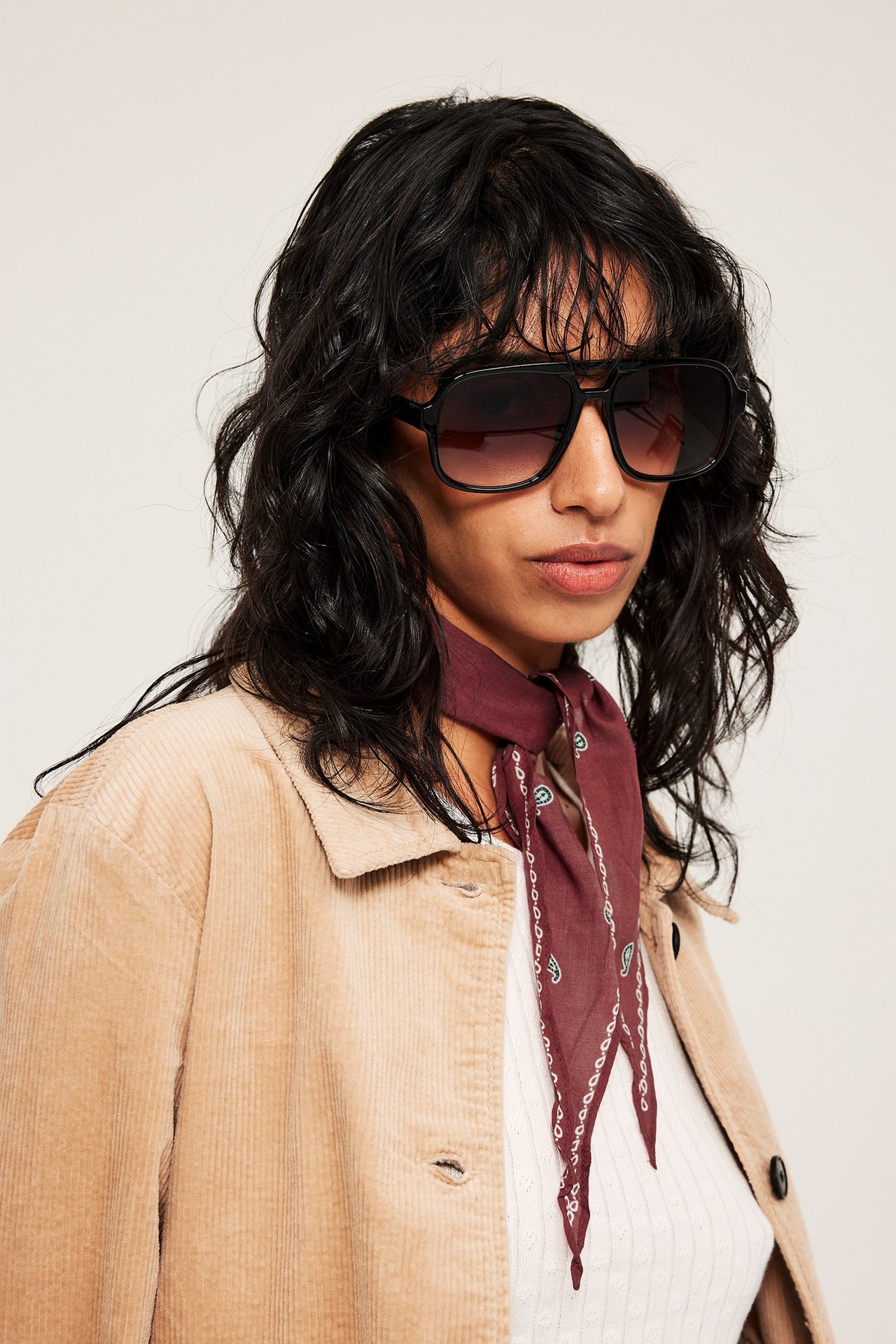 Lähikuva edestä: Nainen punaisessa kuviollisessa rypytetyssä huivissa, mustissa aurinkolaseissa ja beigessä vakosamettitakissa.