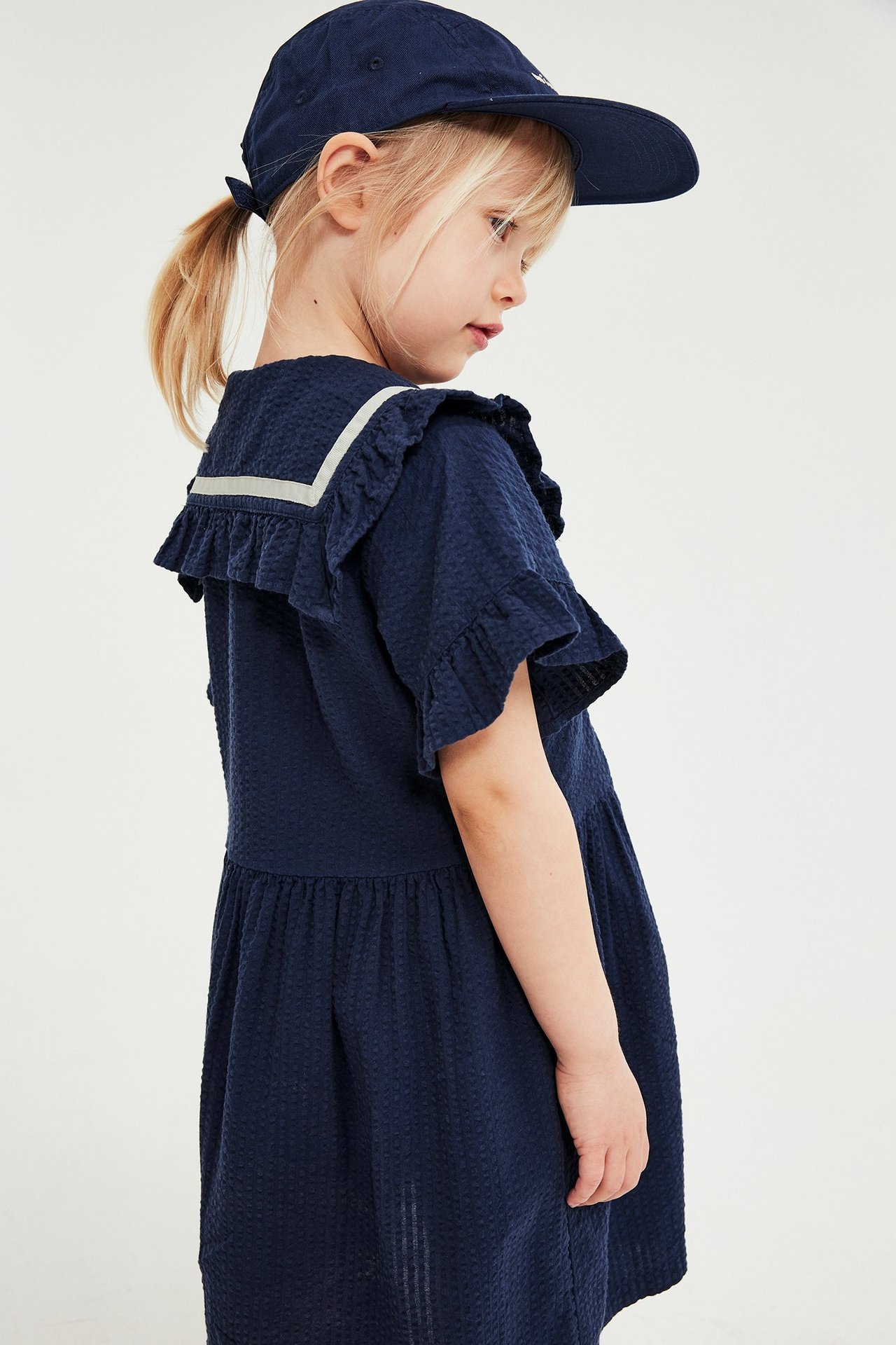Sidevisning av et barn i en marineblå seersucker-kjole med hvitstripet sjømannskrage og en marineblå baseballcaps.