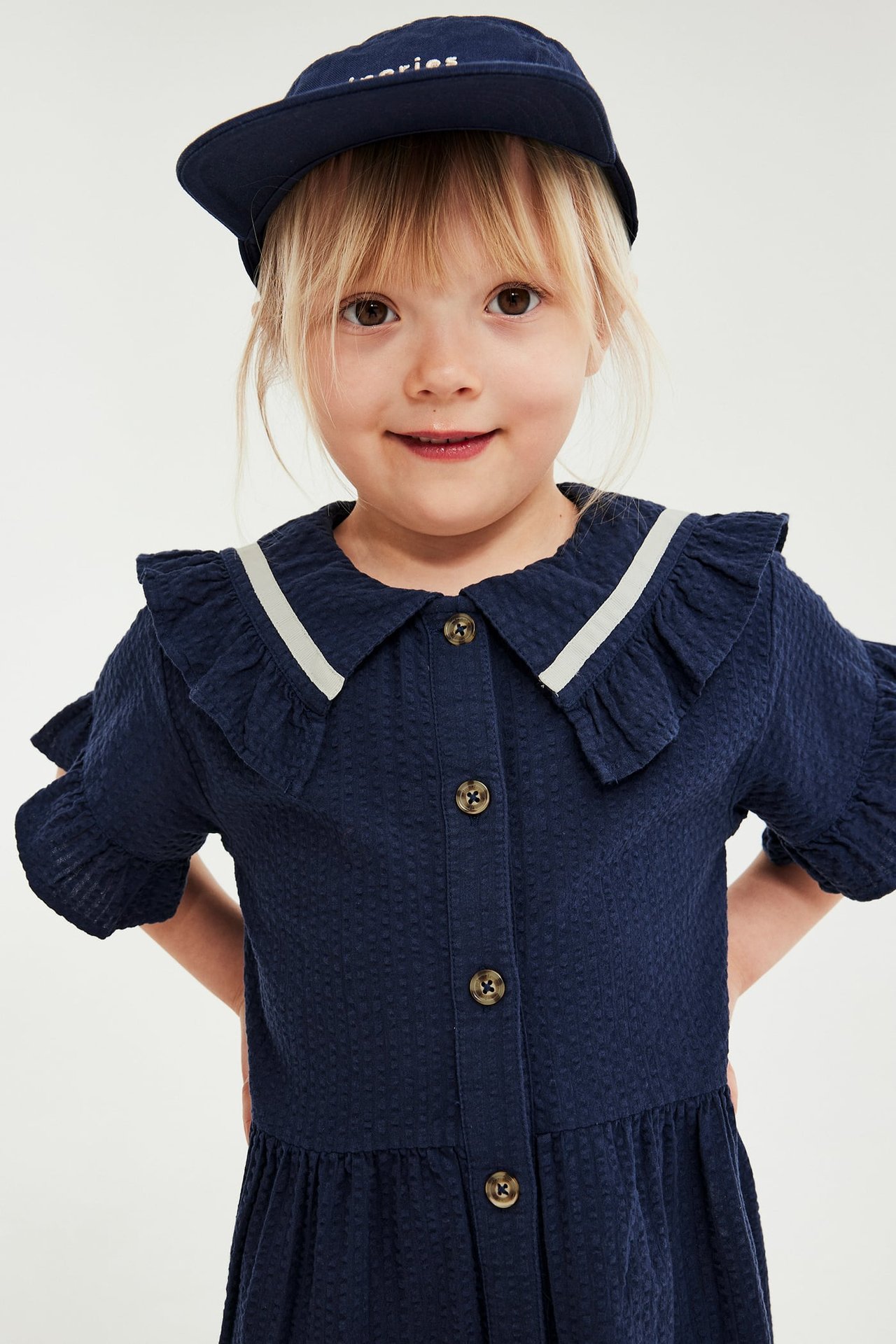 Framifrån, ett barn i mörkblå seersucker-klänning med volanger och knappar, samt en marinblå keps.
