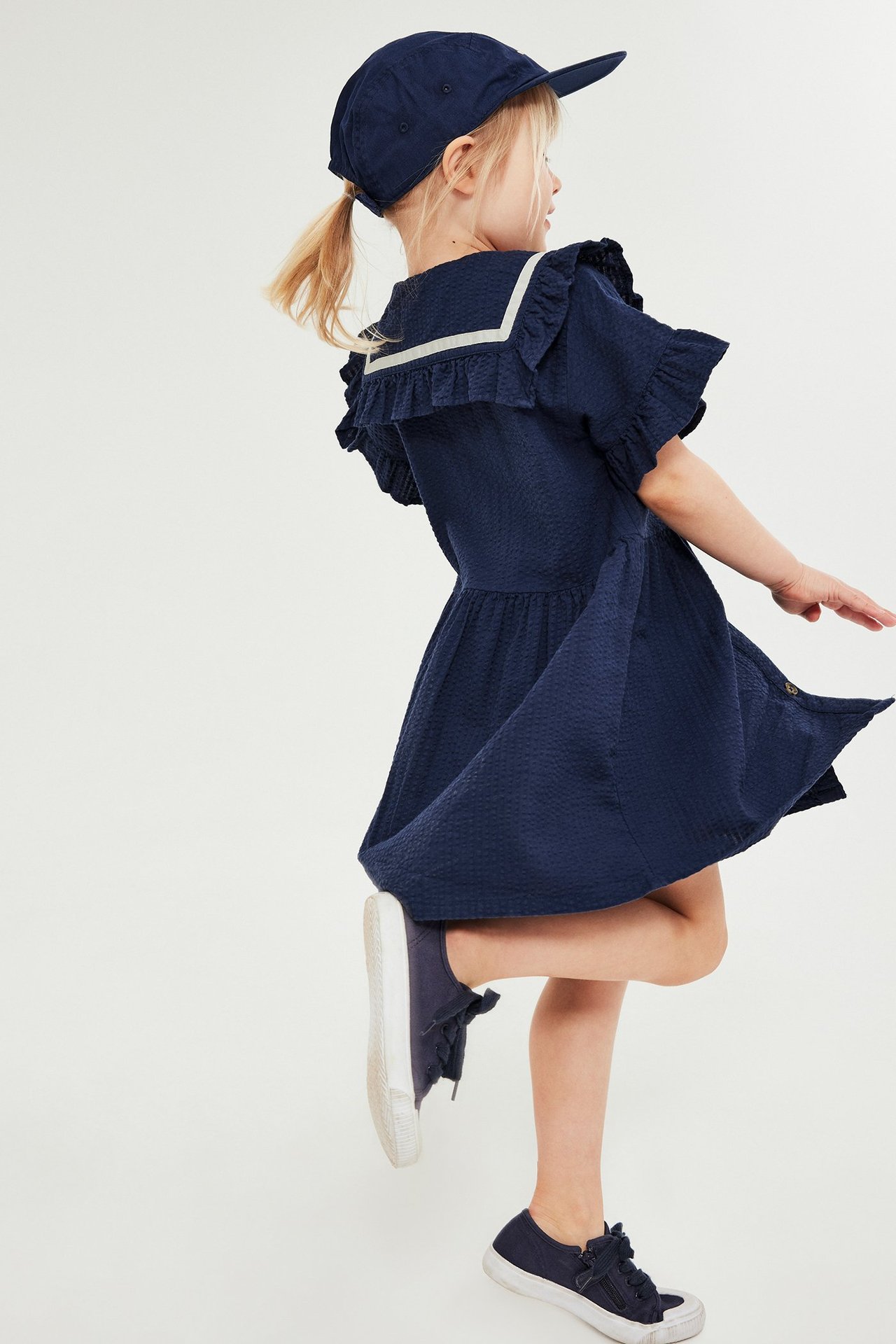 Sivukuva lapsesta yllään tummansininen seersucker-mekko volangikauluksella, lippis ja lenkkarit.