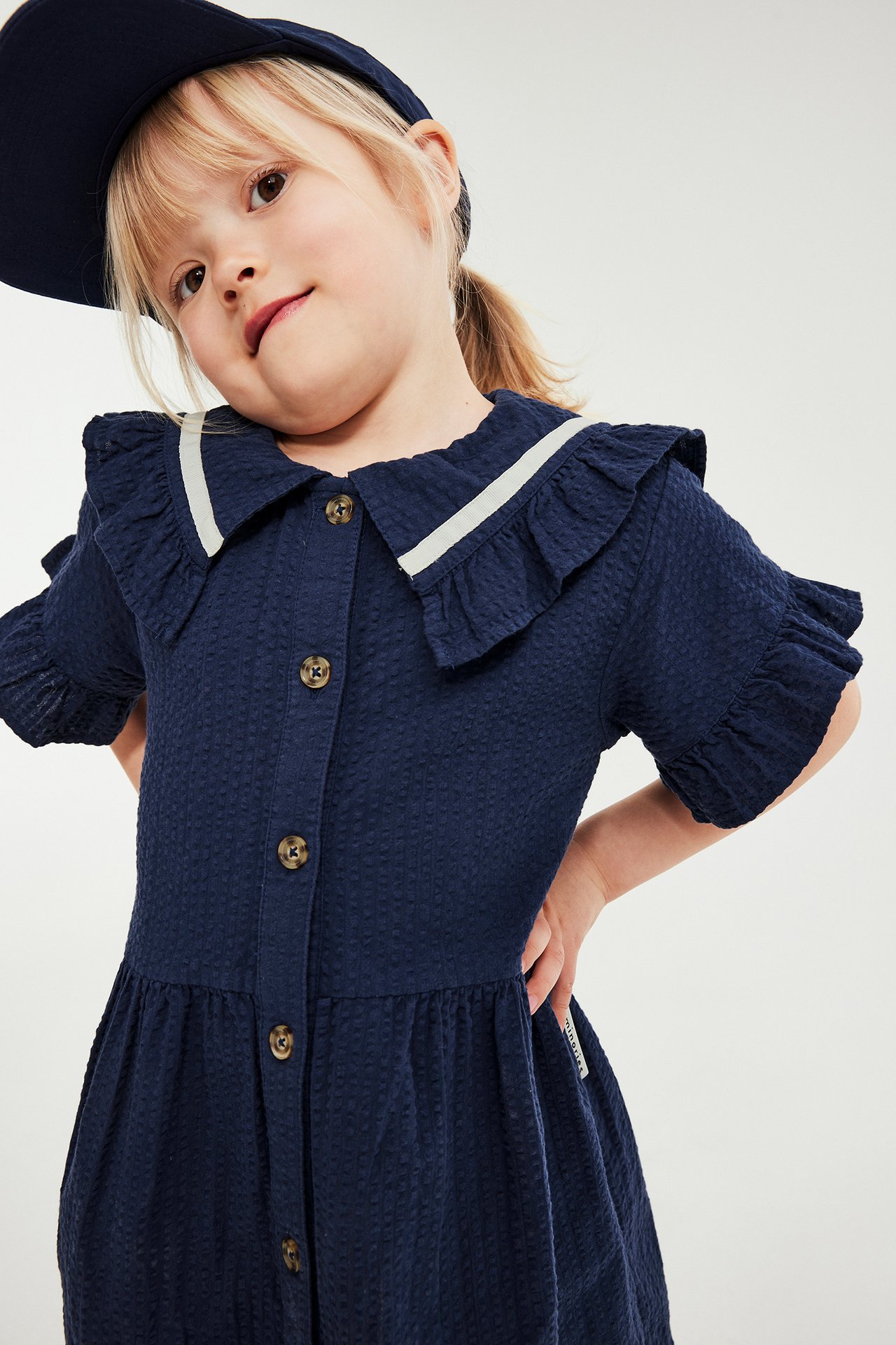Barn i blå strukturert kjole med volangkrage, hvit stripe, knapper foran og blå lue.