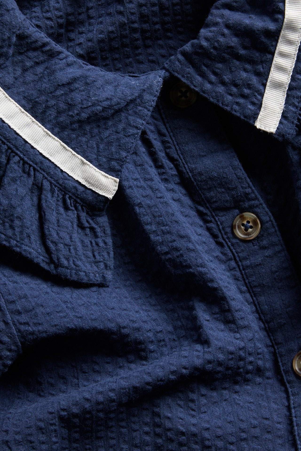 Nærbilde av en marineblå seersucker-kjole med volangkrage, hvit stripet kant og brune knapper foran.