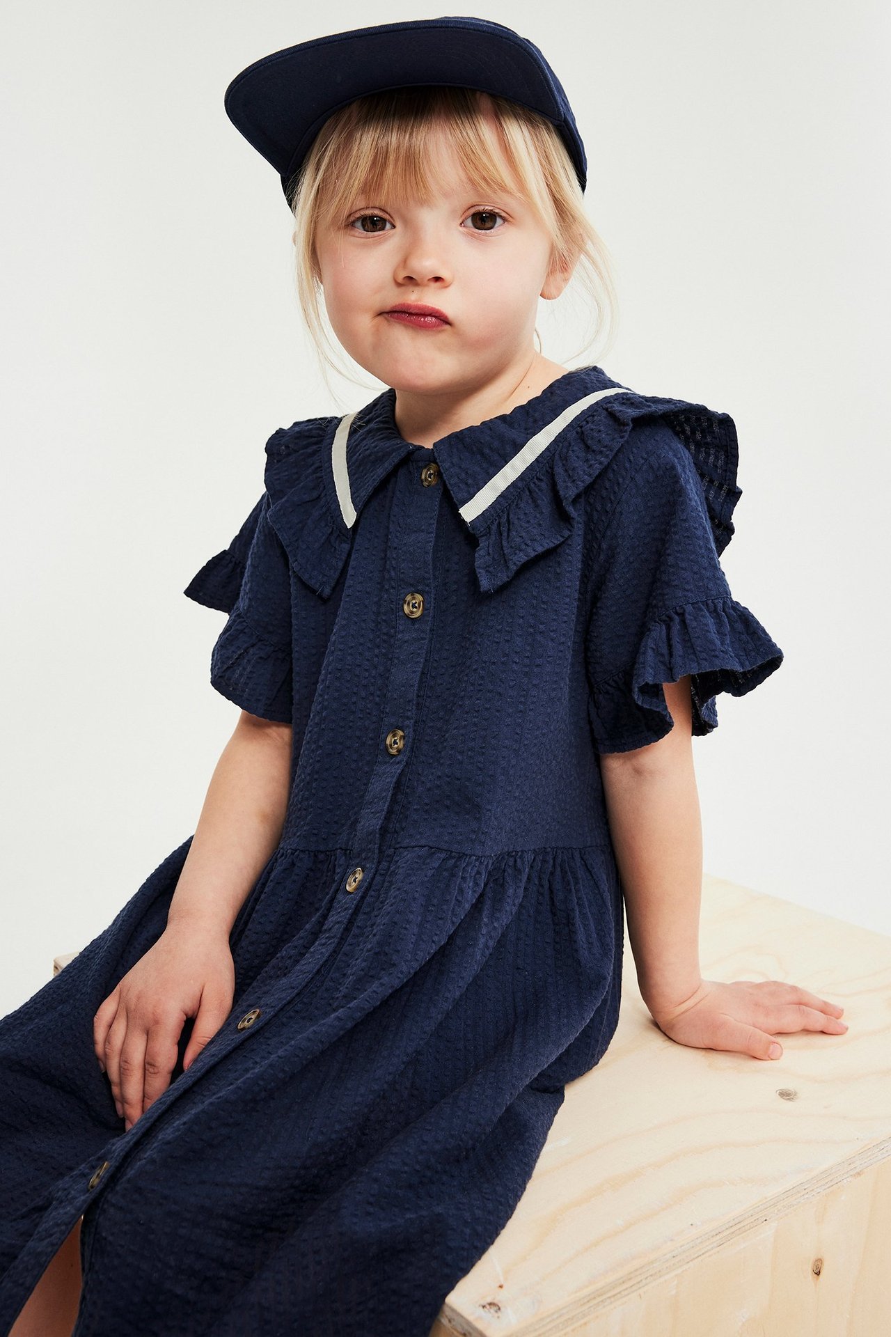 Framifrån, ett barn i en marinblå seersuckerklänning med volanger, vitkantad krage och en matchande keps.