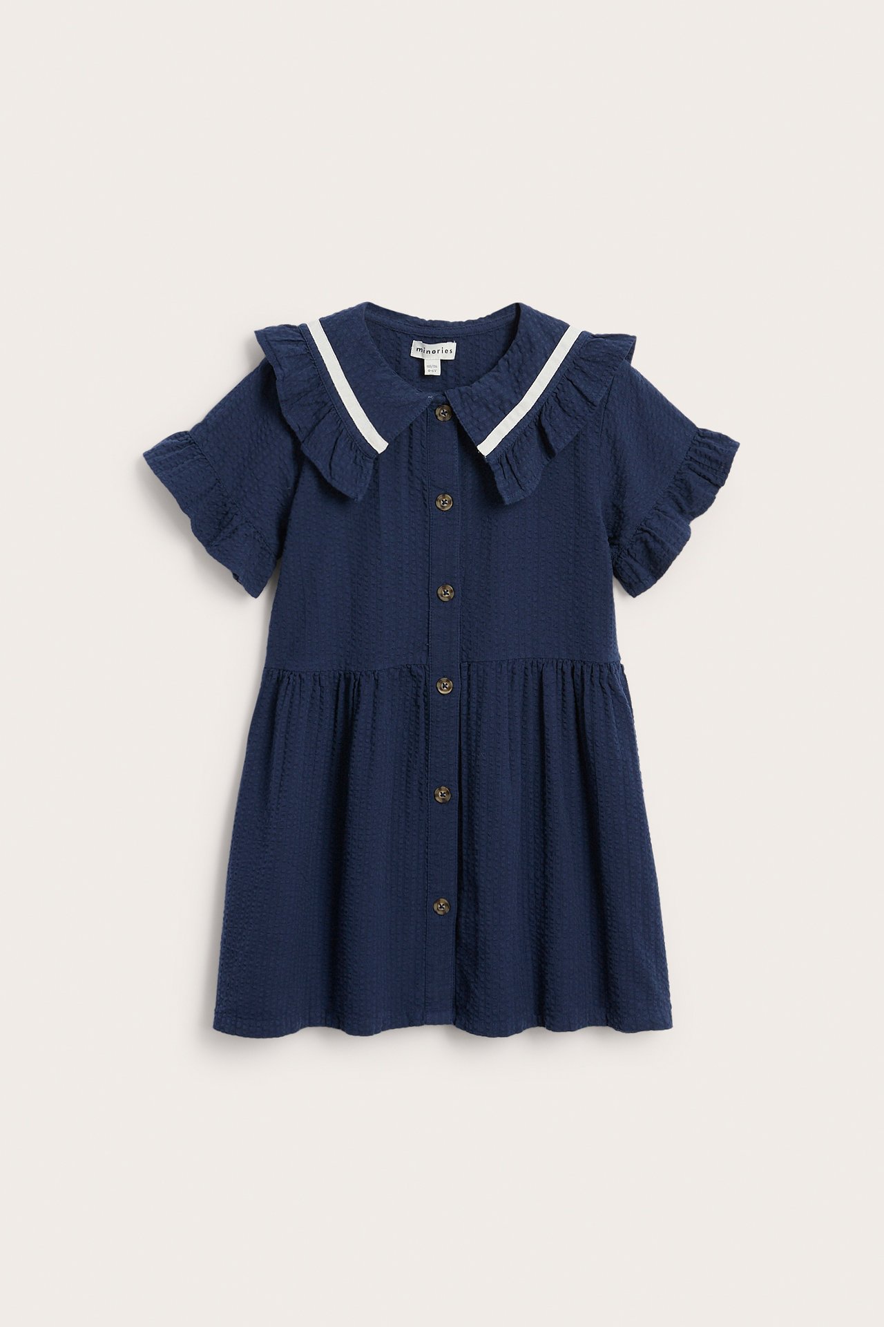 Frontvisning av en mørkeblå seersucker-kjole for barn med volangkrage, hvit kant og knapper foran.