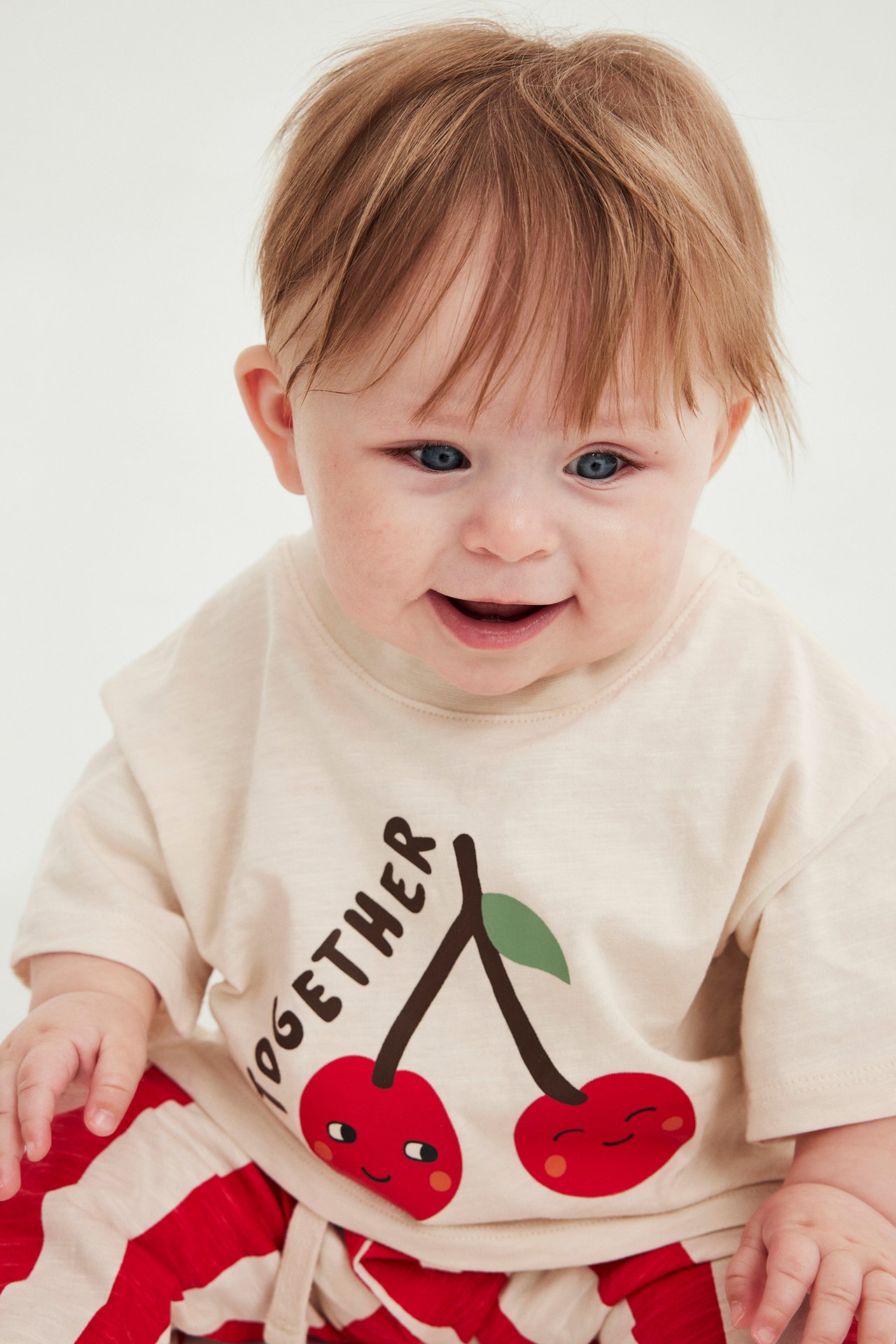 Leende baby i beige bomulls-T-shirt med körsbärstryck och rödrandiga bomullsbyxor, framifrån.