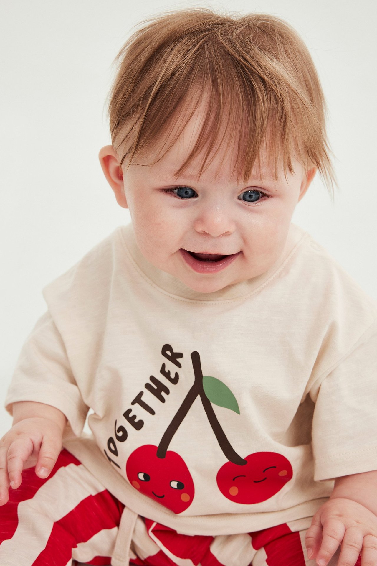 Hymyilevä vauva yllään beige puuvillainen T-paita kirsikkaprintillä ja punaraitaiset puuvillahousut, edestä.