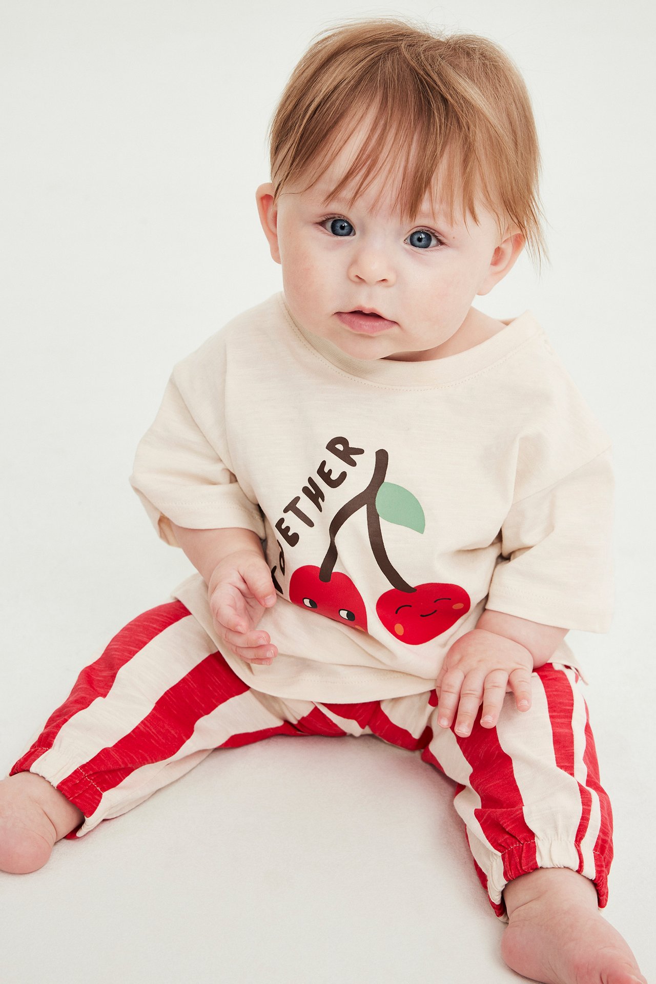 Framifrån, baby i offwhite T-shirt med körsbärstryck och rödrandiga joggingbyxor.