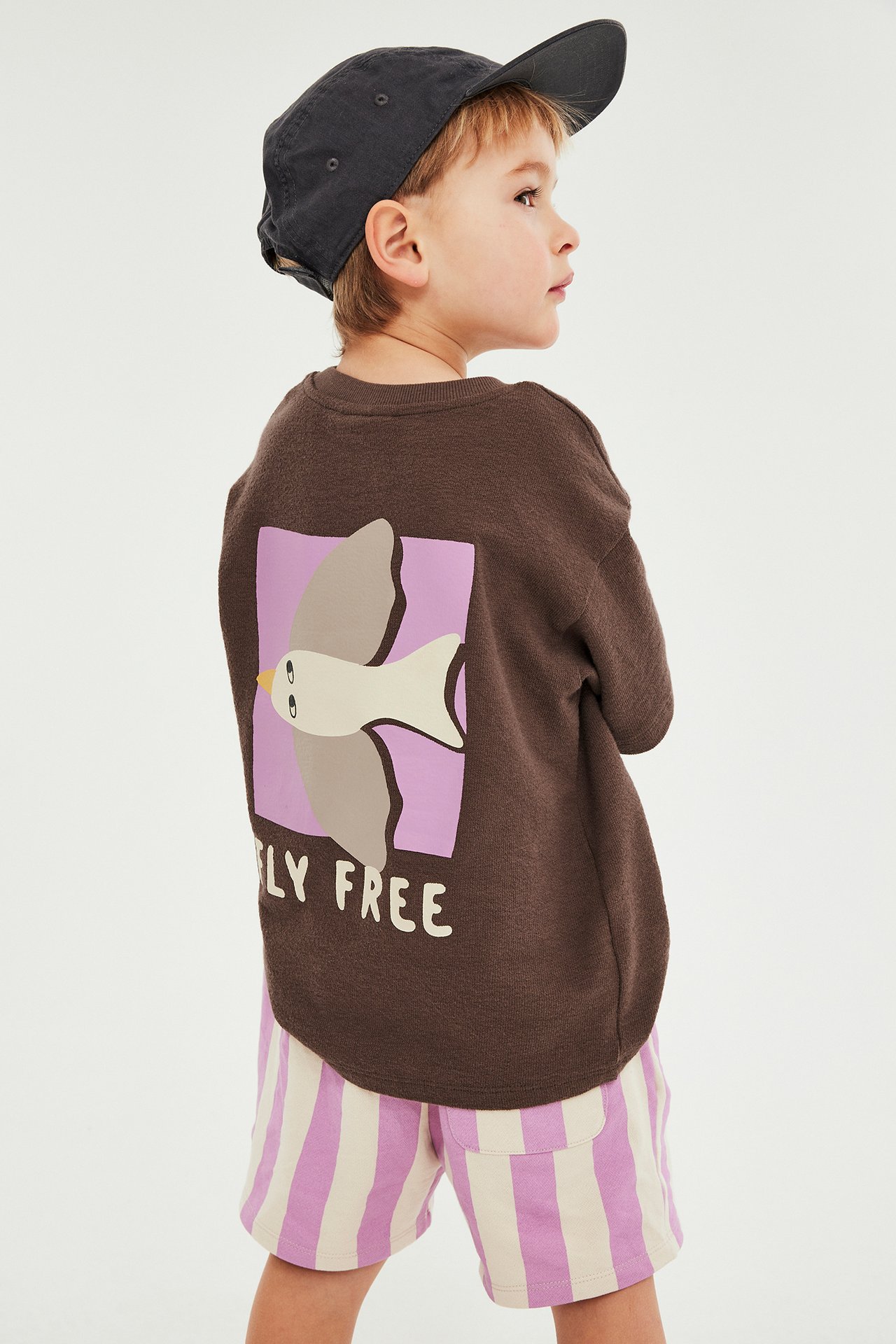 Ryggvisning barn: Brun kortermet t-skjorte med lilla fugletrykk, lyserosa stripete shorts og mørkegrå caps.