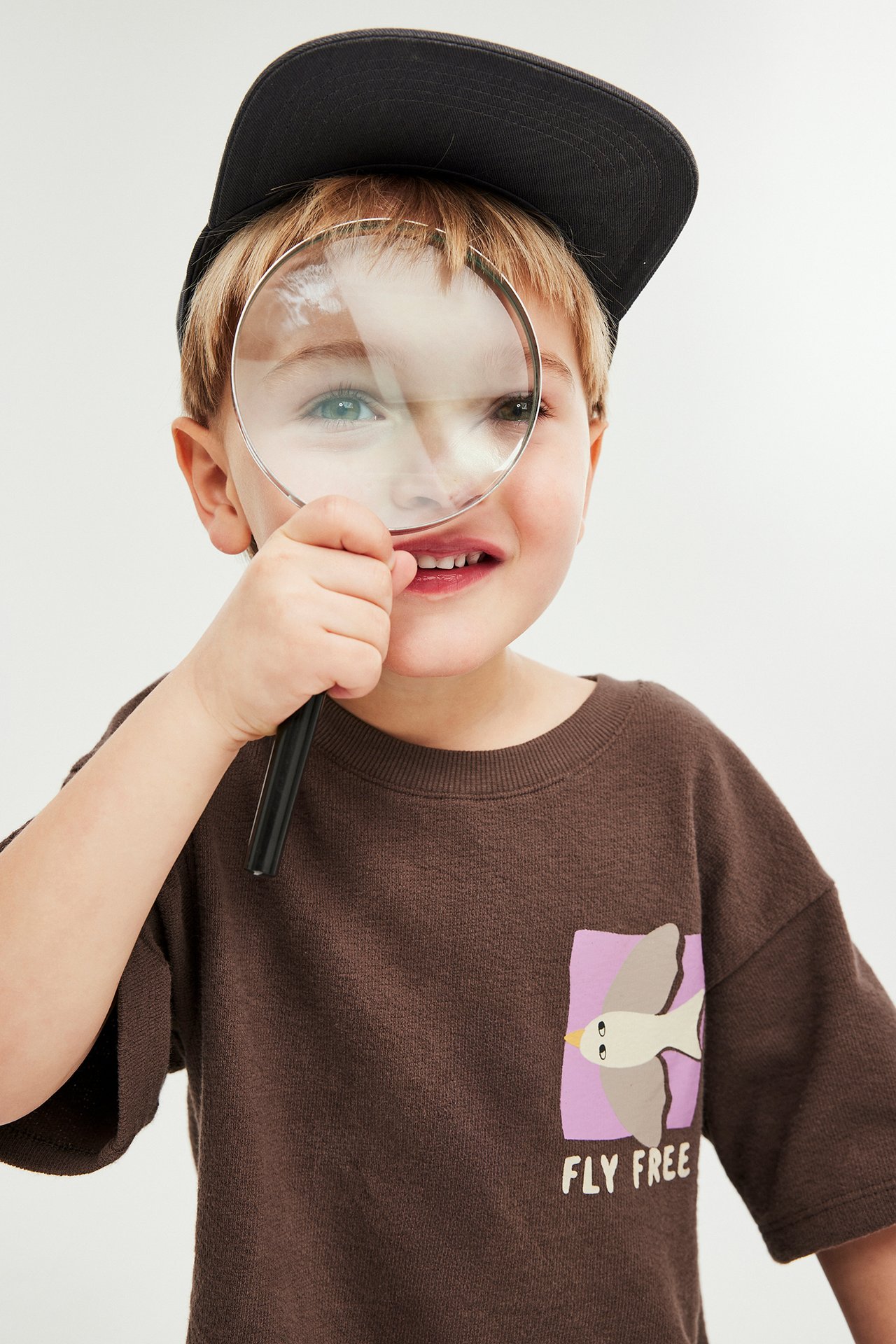 Lapsi ruskeassa puuvillaisessa t-paidassa lintupainatuksella, musta lippalakki, pitäen suurennuslasia.