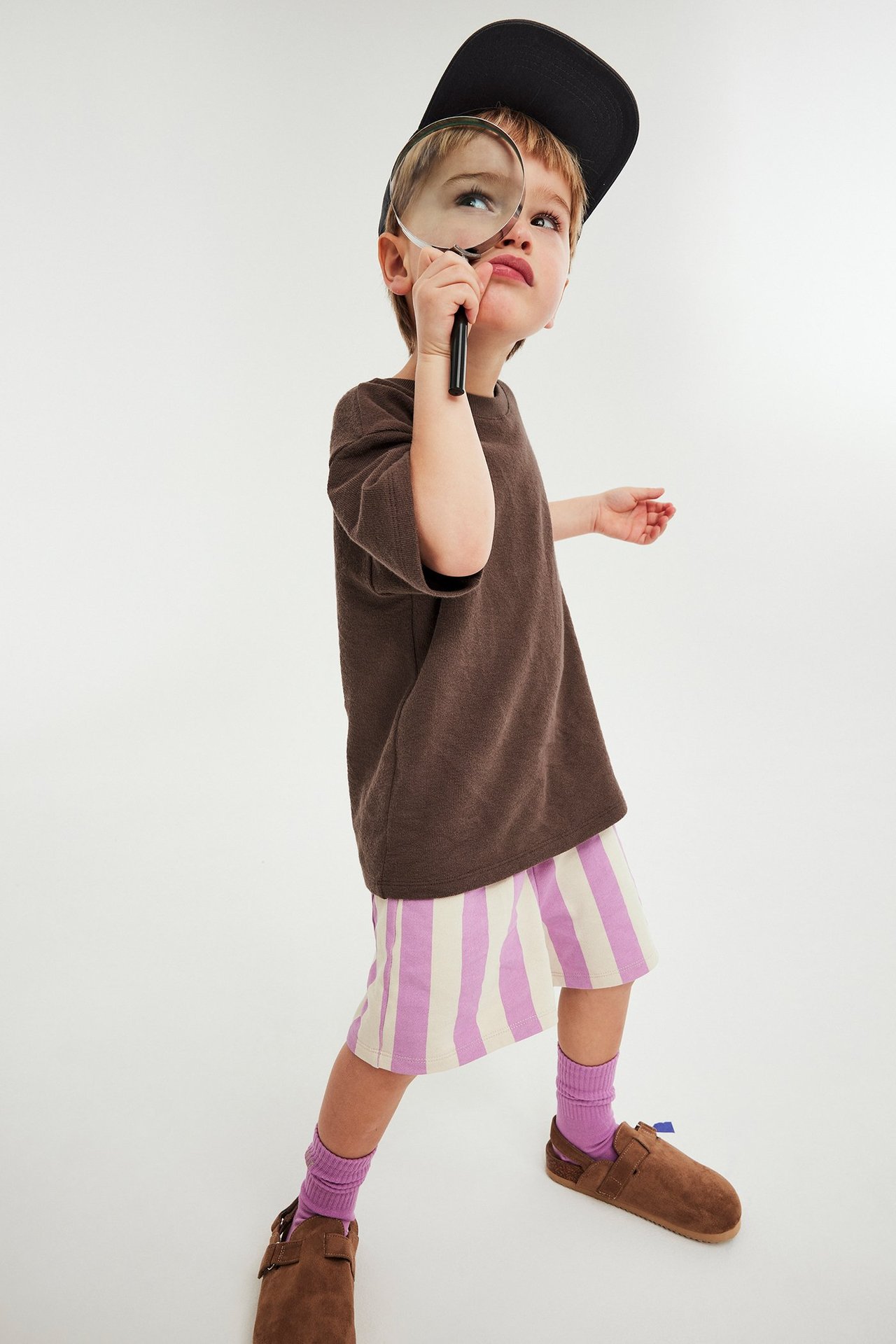 Forfra: Barn i brun t-skjorte, lilla stripete shorts, lilla sokker, brune tresko, svart caps, holder forstørrelsesglass.