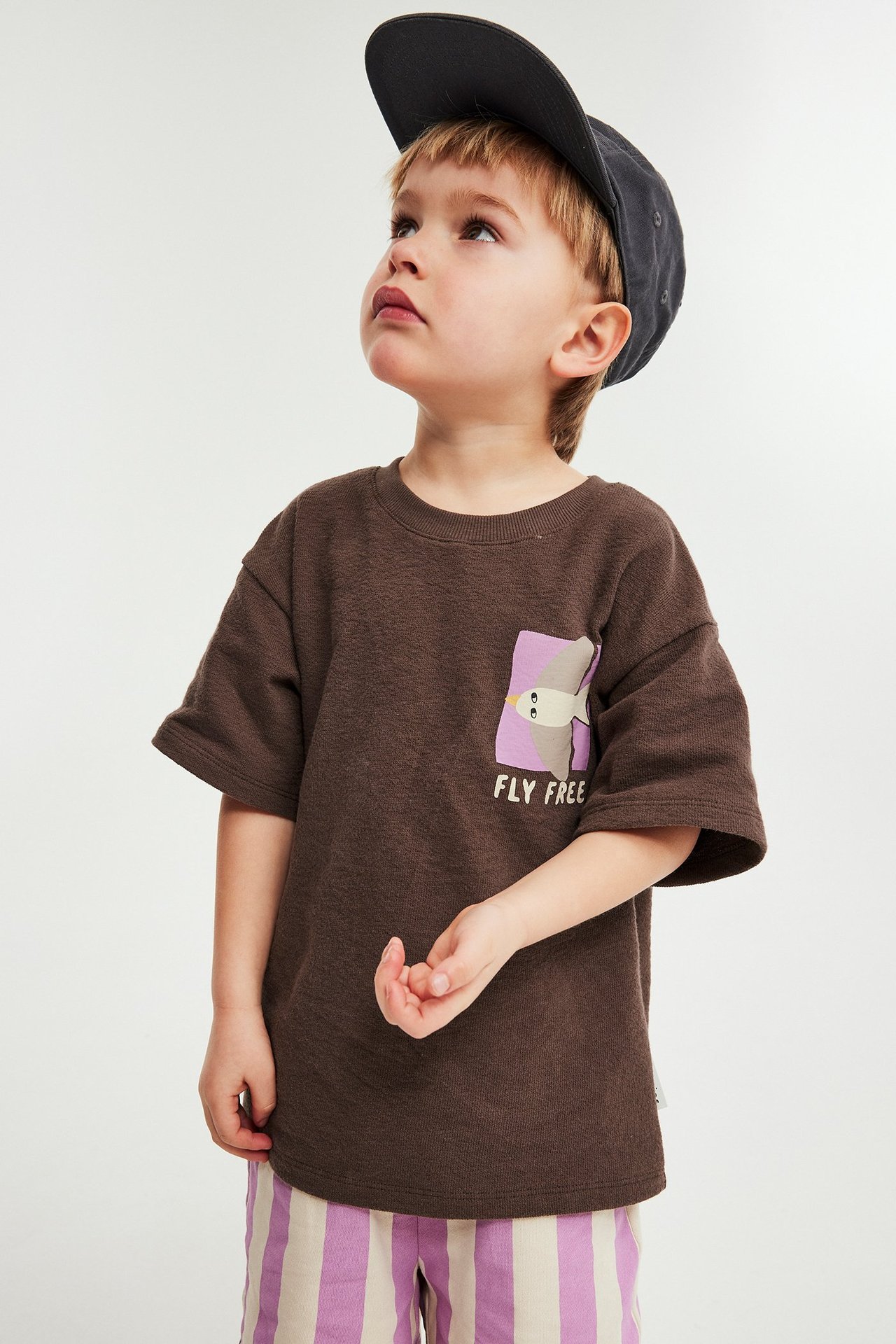 Forfra: Et barn har på seg en brun bomulls-t-skjorte med fugletrykk, stripete shorts og mørk caps.