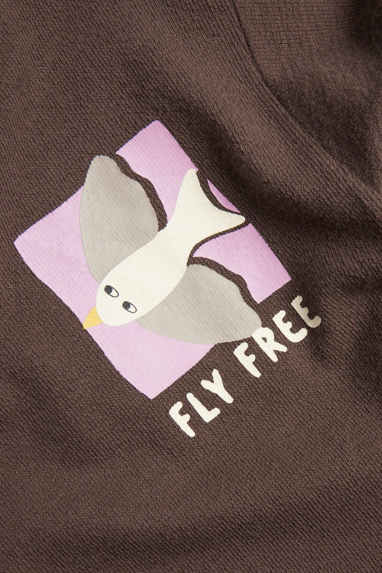 Zbliżenie na brązowy bawełniany T-shirt dziecięcy z nadrukiem białego ptaka i napisem "FLY FREE".