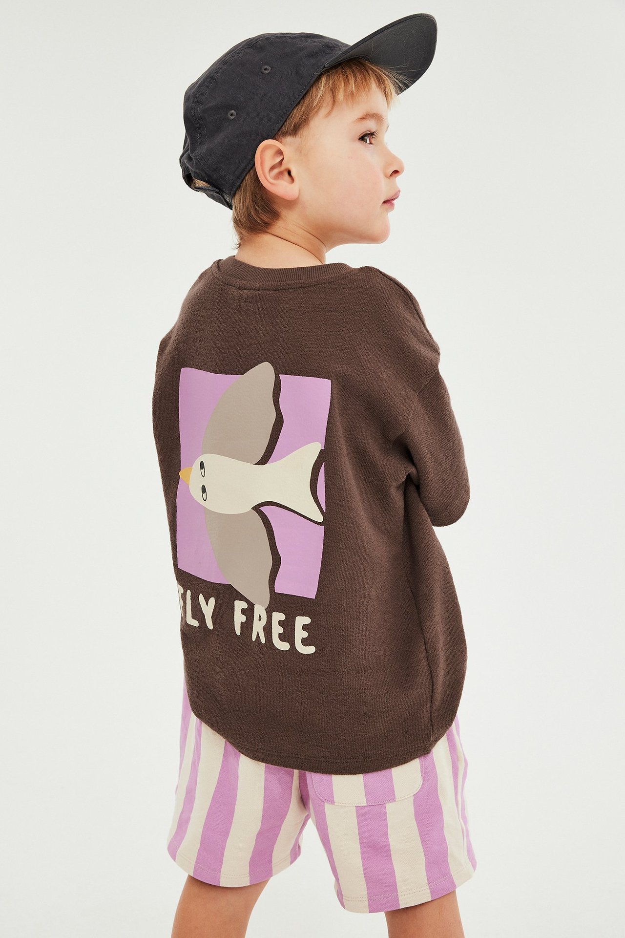 Widok z tyłu: brązowy T-shirt z krótkim rękawem z fioletowym ptakiem, różowe szorty w paski, ciemnoszara czapka.