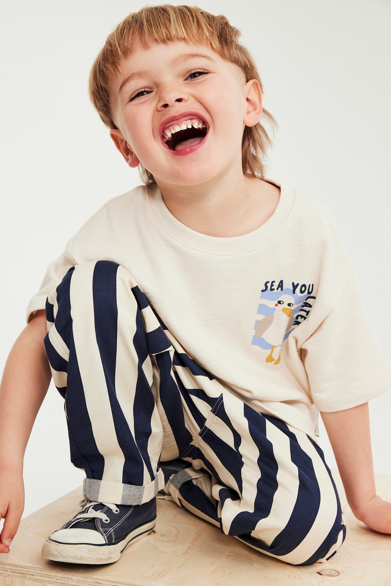 Framifrån. Ett barn i ljusbeige bomulls-t-shirt med måstryck och marinblårandiga byxor.