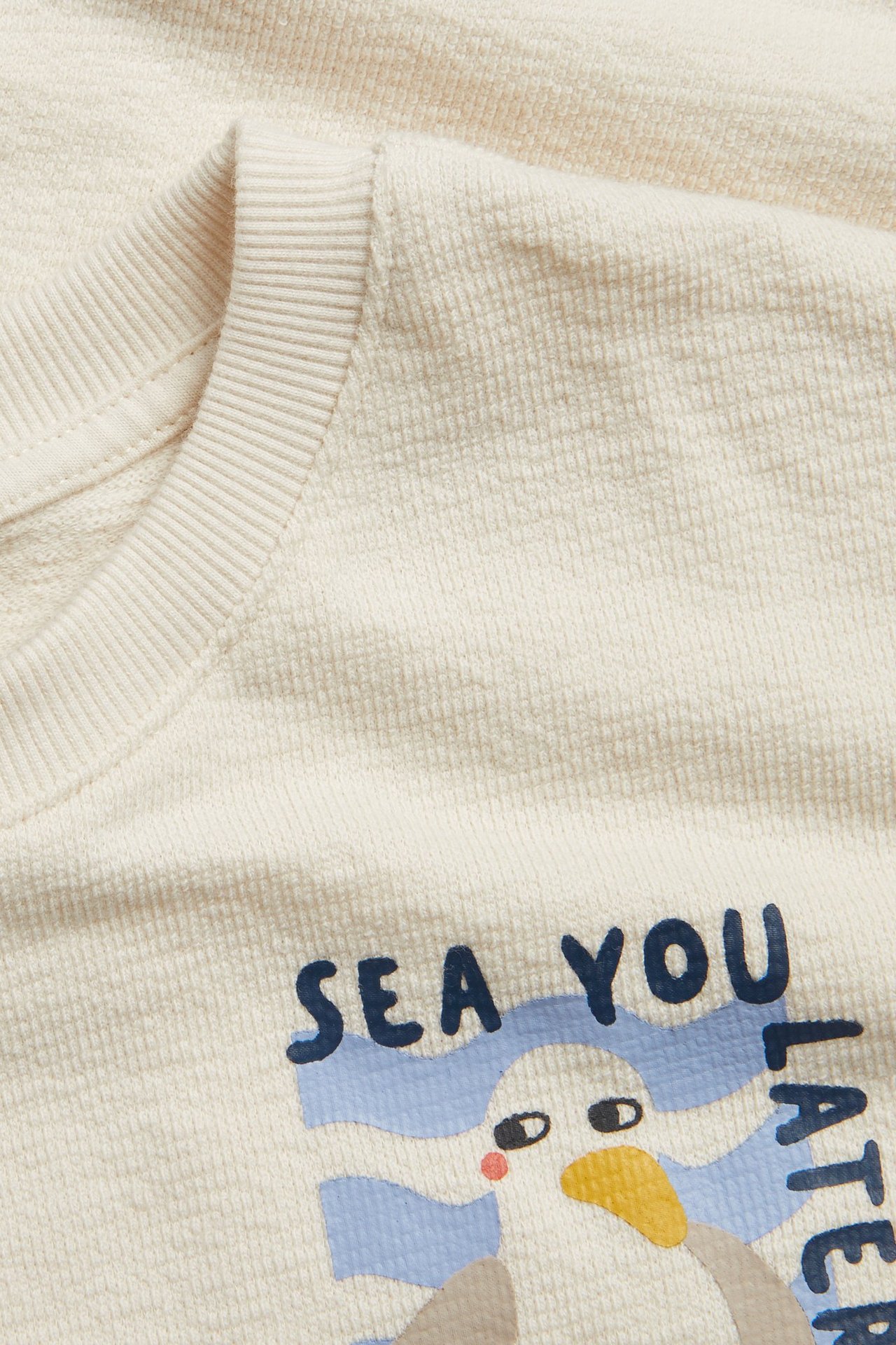 Nærbilde forfra av en lys beige kortermet T-skjorte i bomull for barn med "SEA YOU LATER"-trykk og måkegrafikk.