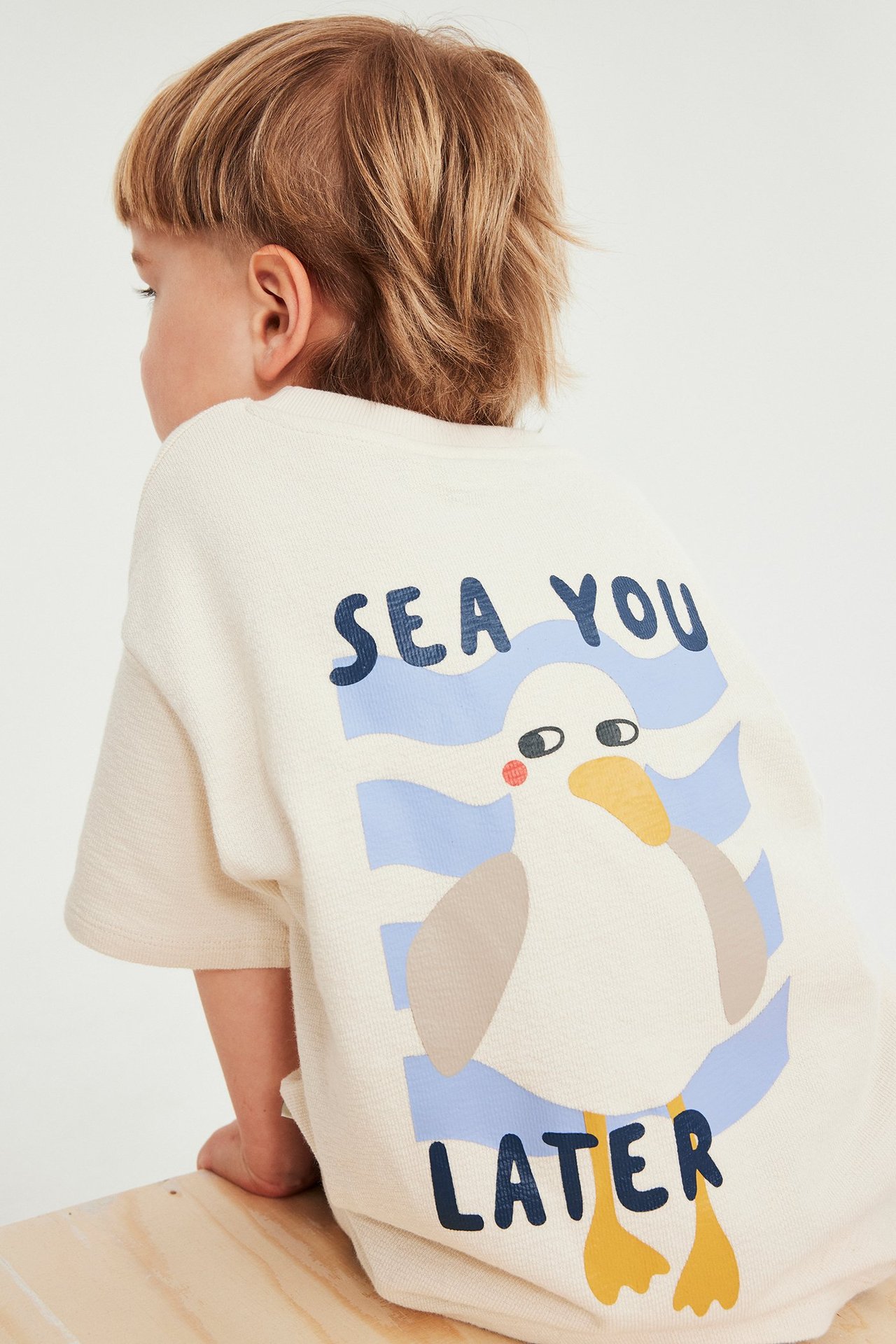 Ryggvy av ett barn i en ljusbeige bomulls-t-shirt med sjöfågelprint och texten "SEA YOU LATER".