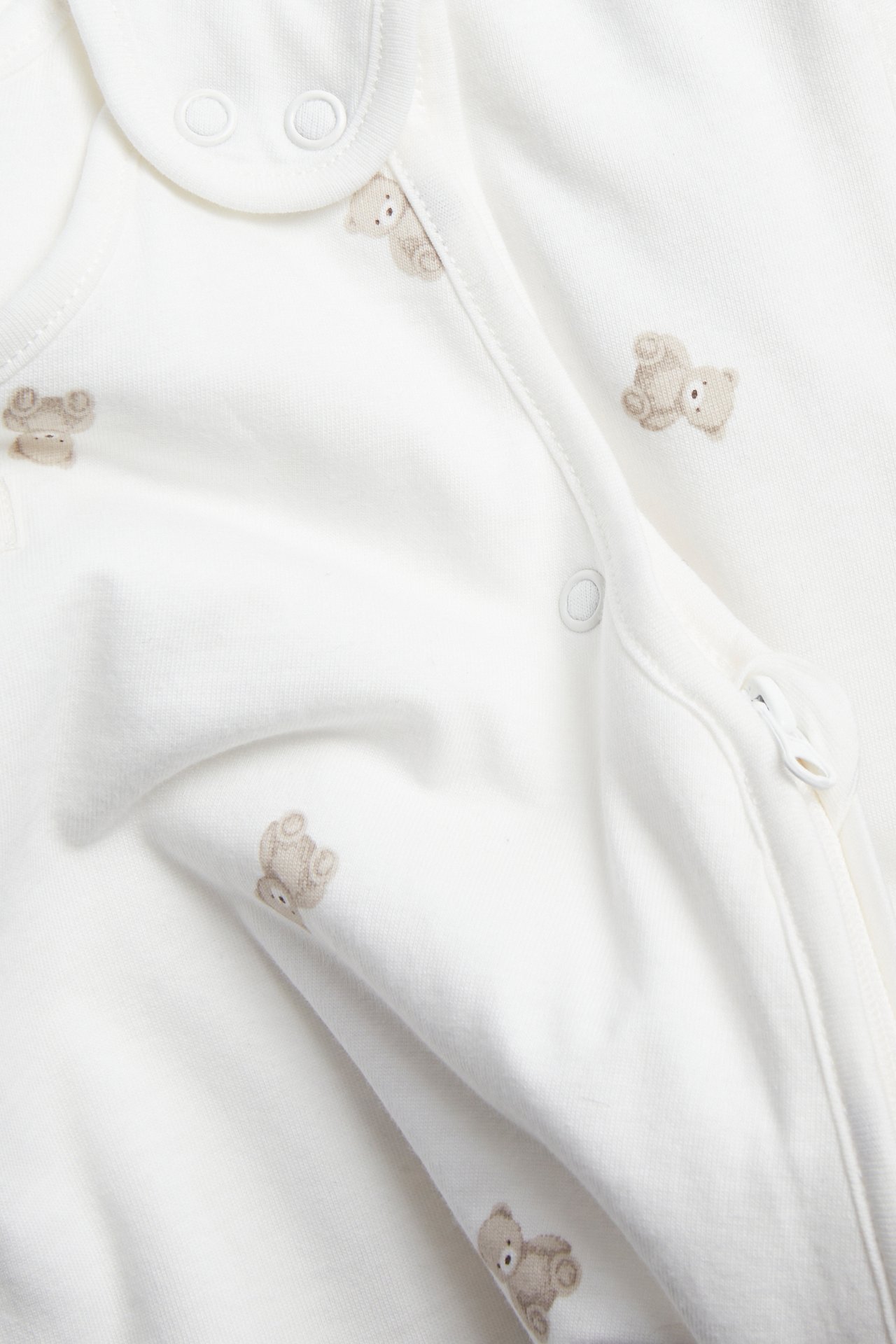 Lähikuva luonnonvalkoisesta vauvan unipussista, jossa vaaleanruskea karhukuvio, painonapit ja kaksisuuntainen vetoketju.