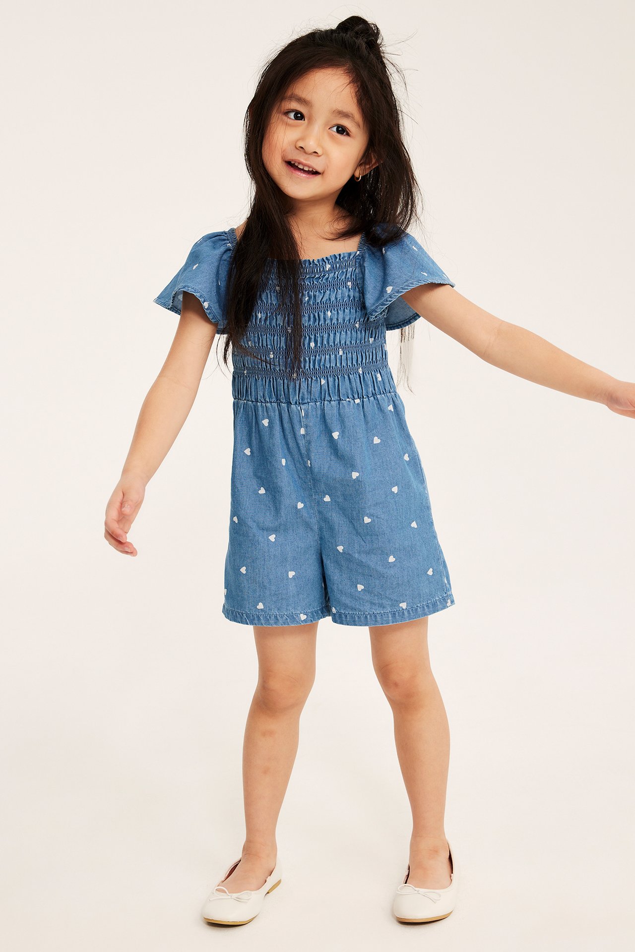 Frontbilde: Jente i lys blå denimplaysuit med hjertemønster, smocktopp og hvite ballerinasko.