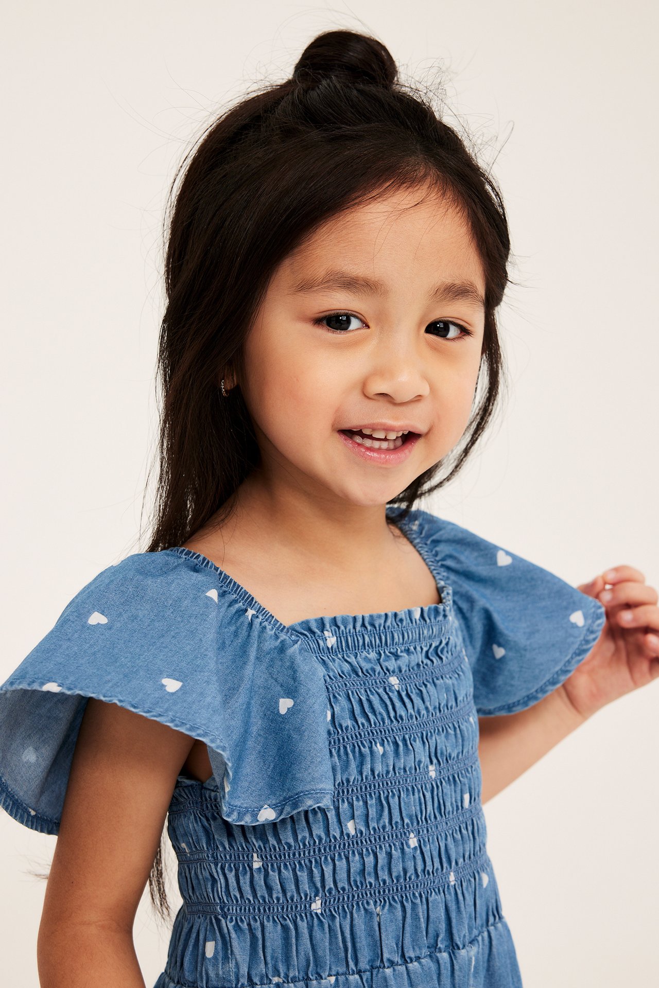 Från framsidan: Barn i ljusblå denim-playsuit med vitt hjärtmönster, volangärmar, smock upptill.