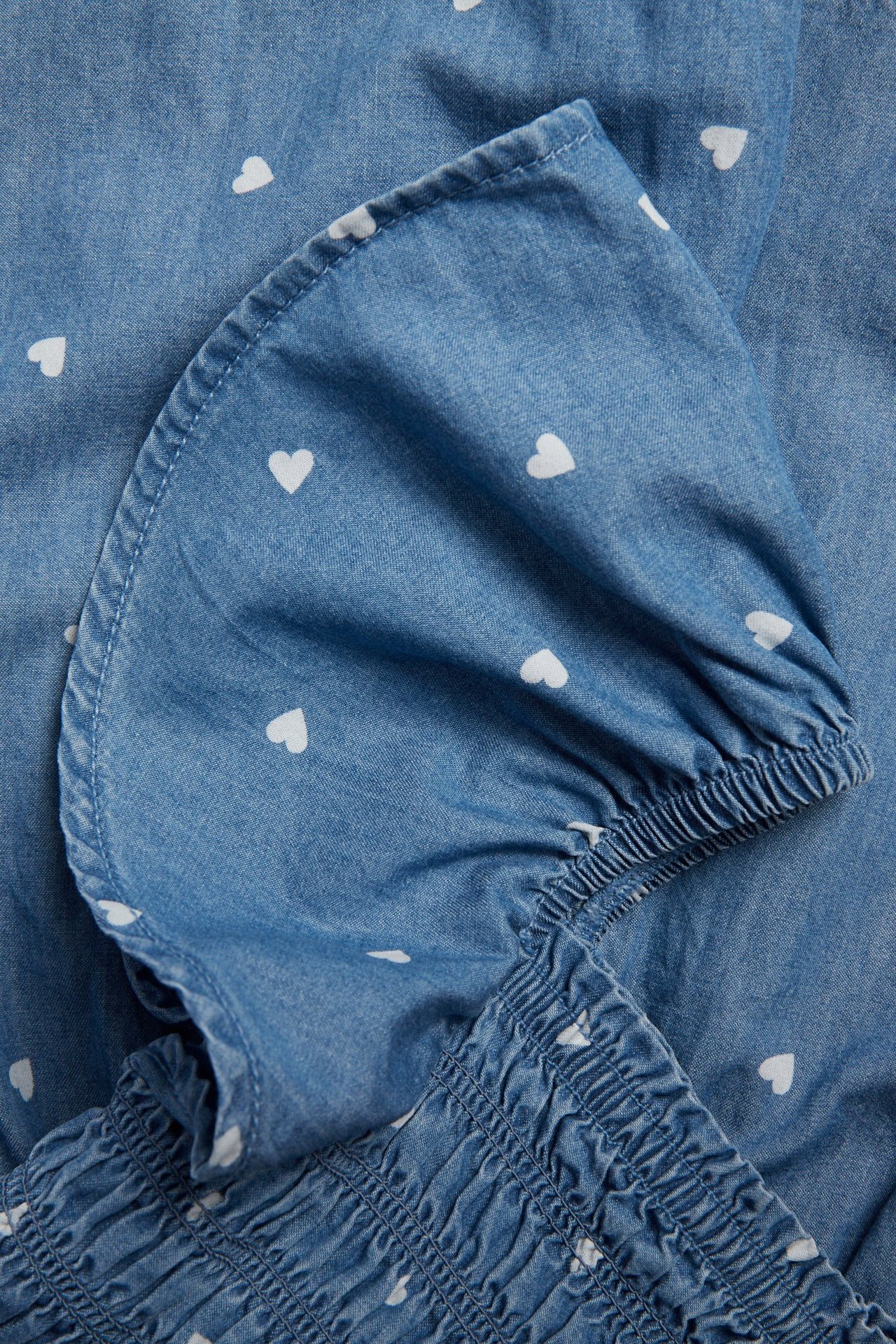 Nærbilde av en lyseblå denimplaysuit med hvitt hjertemønster, smocket topp og elastisk volangerm.