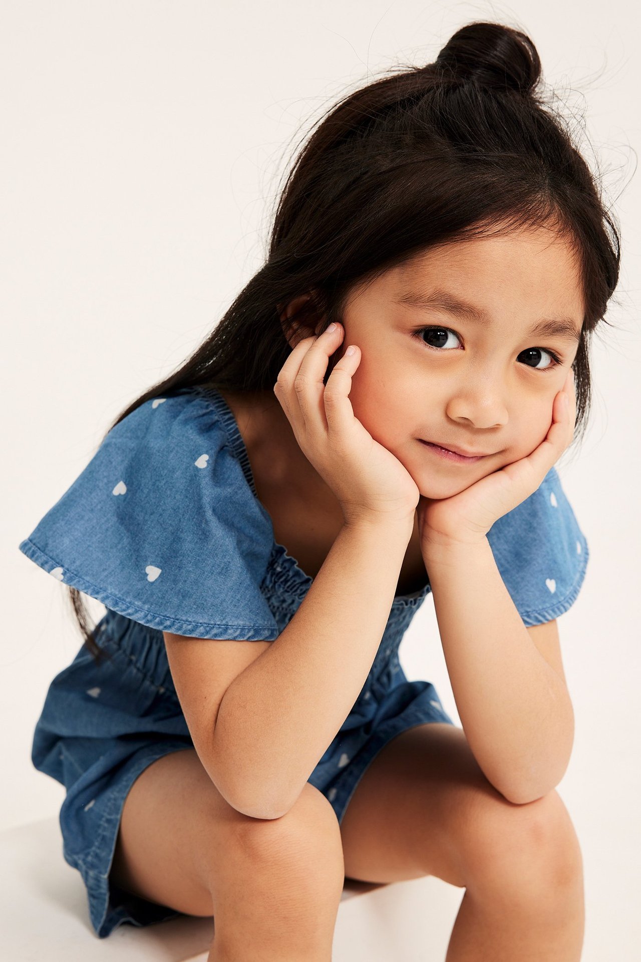 Edestäpäin, lapsi yllään vaaleansininen denim-playsuit valkoisella sydänkuviolla, smokkirypytyksellä ja röyhelöhihoilla.