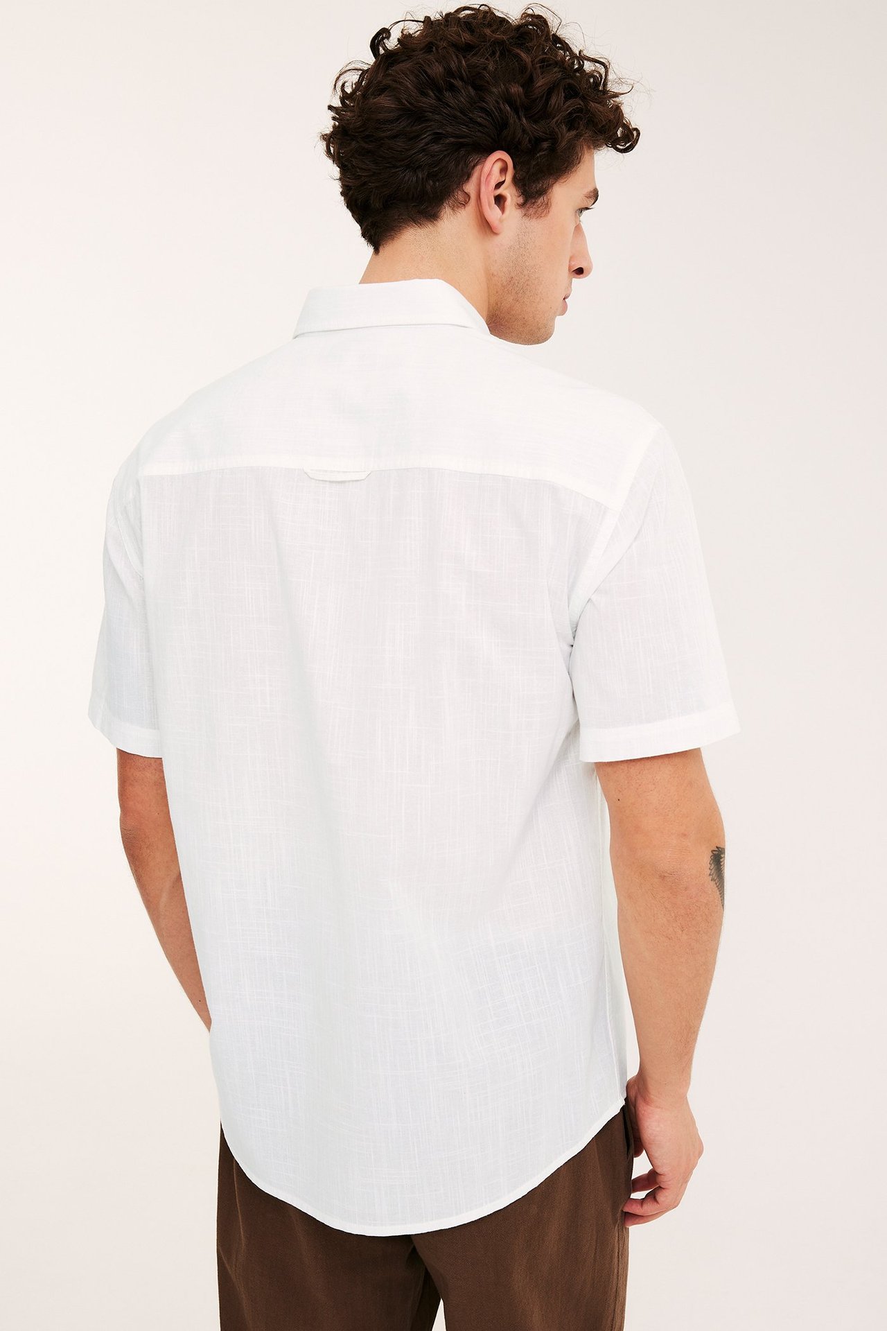Widok z tyłu mężczyzny w białej, teksturowanej, krótko-rękawowej koszuli z mieszanki bawełny i brązowych spodniach.