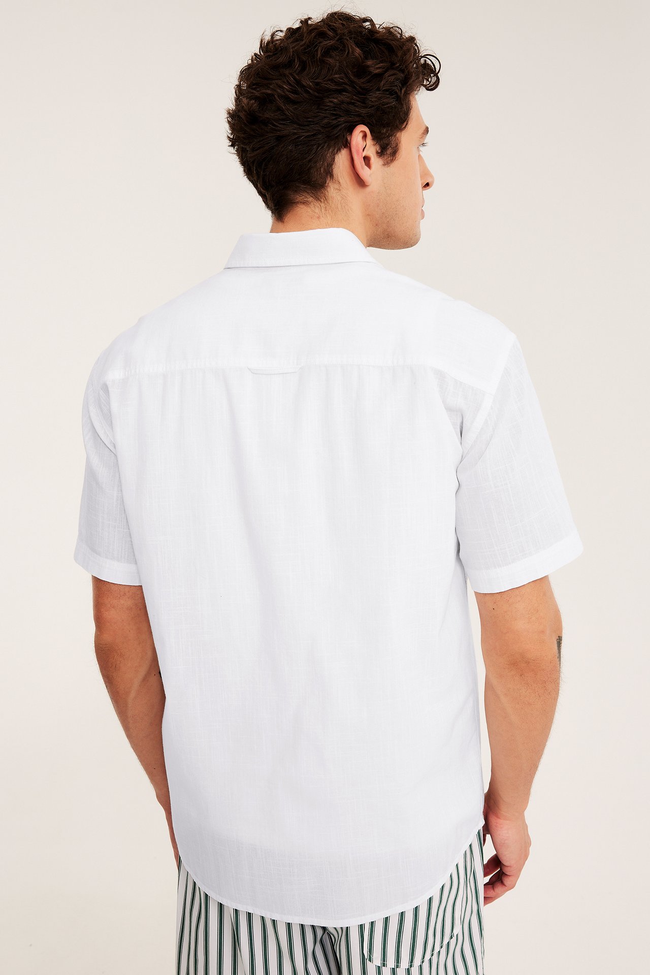 Takanäkymä miehestä valkoisessa, normaalimittaisessa, lyhythihaisessa kuvioidussa paidassa ja raidallisissa shortseissa.