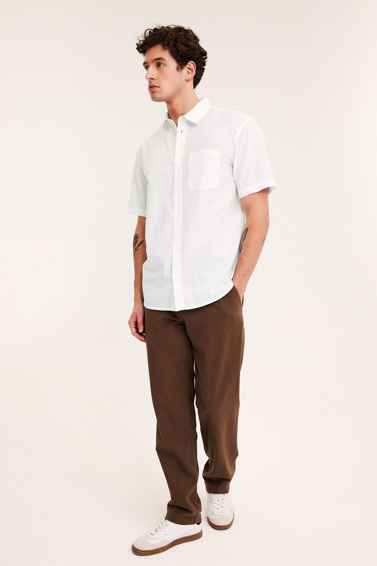 Man i sidoprofil bär vit regular fit kortärmad strukturerad skjorta, bruna byxor och vita sneakers.
