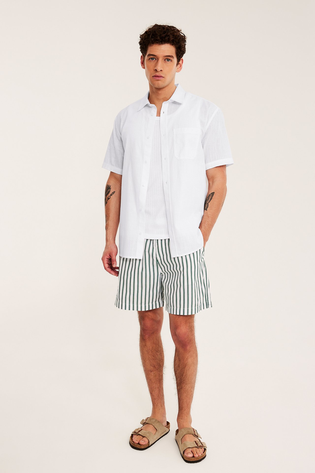 Framifrån: Man bär vit kortärmad regular fit skjorta, grönrandiga shorts och beige sandaler.