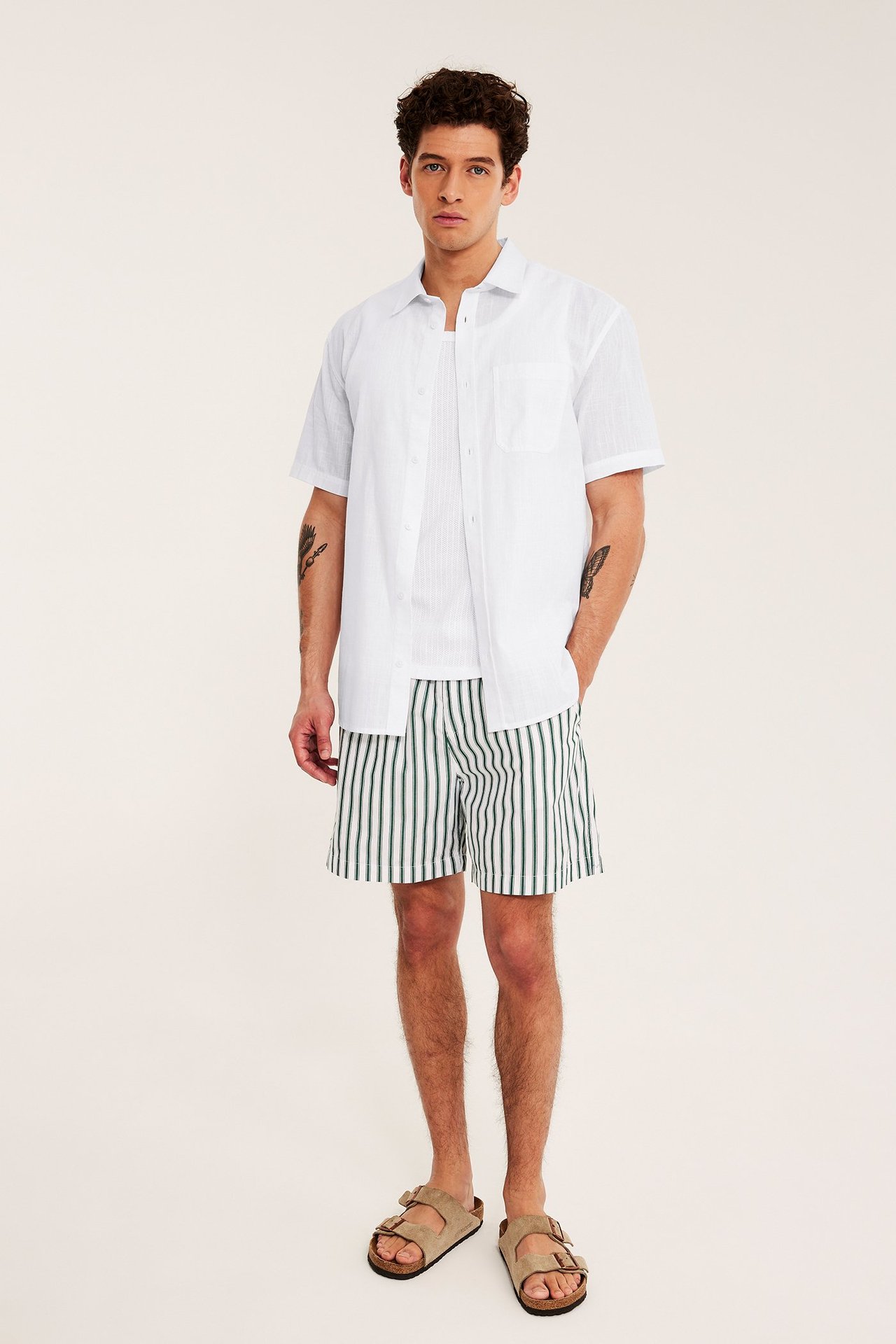Widok z przodu: Mężczyzna ma na sobie białą koszulę regular fit z krótkim rękawem, zielone spodenki w paski i beżowe sandały.