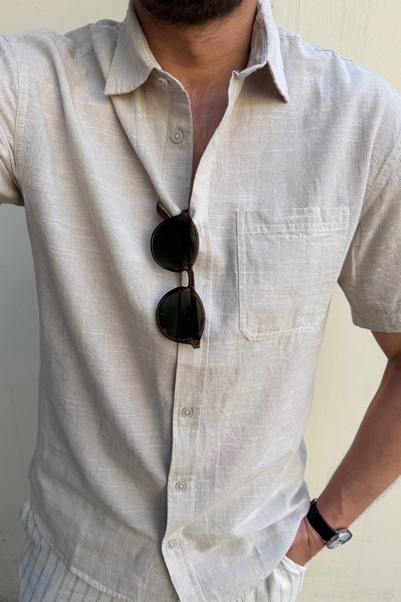 Närbild framifrån: man i beige kortärmad regular fit skjorta, bröstficka, mörka solglasögon hängandes.