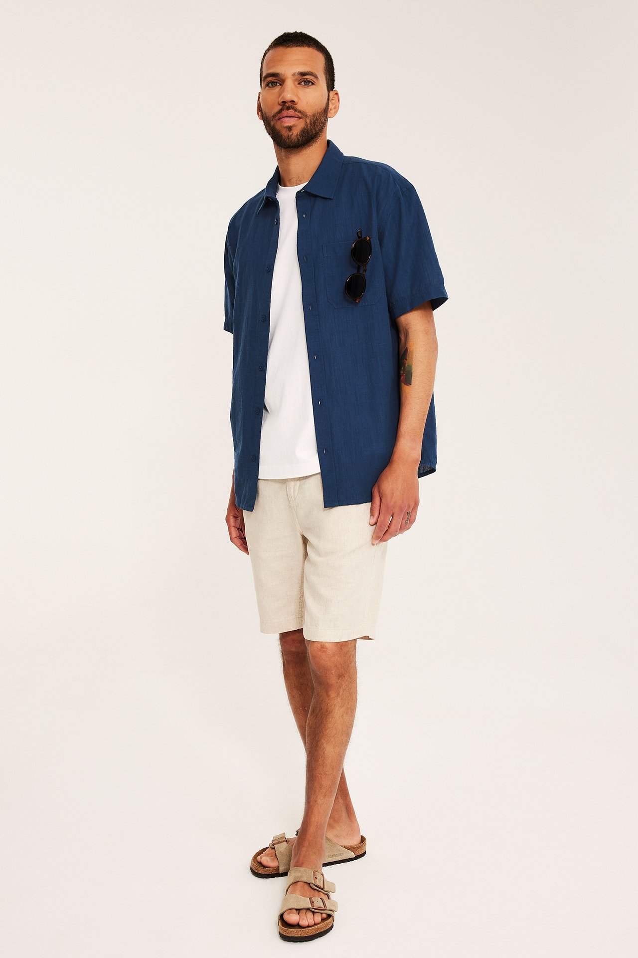 Framifrån: Man i blå strukturerad kortärmad skjorta, vit t-shirt, beiga shorts och sandaler. Solglasögon i fickan.
