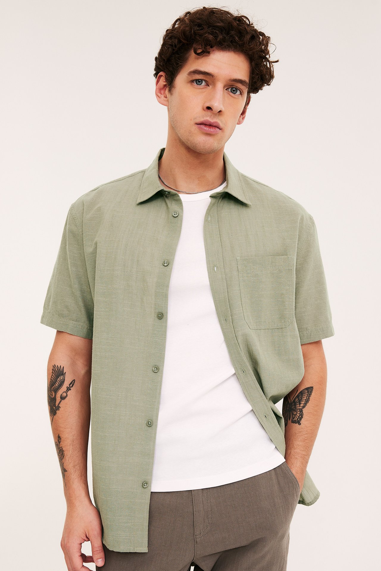 Framifrån: Man bär en khakigrön strukturerad kortärmad skjorta över en vit t-shirt och mörka byxor.