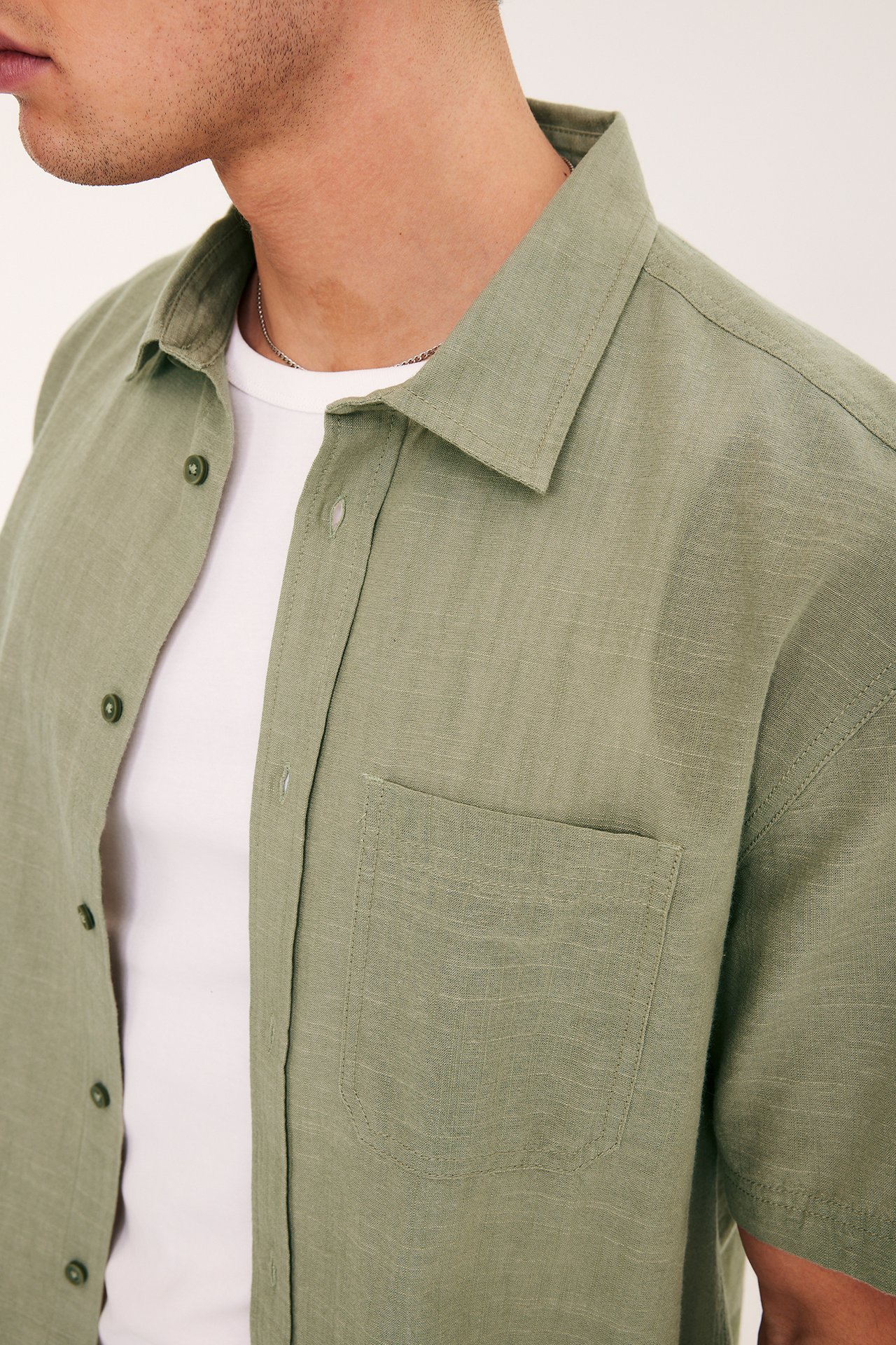 Mężczyzna w otwartej khakizielonej koszuli regular fit z krótkim rękawem i kieszenią, na białym T-shircie.