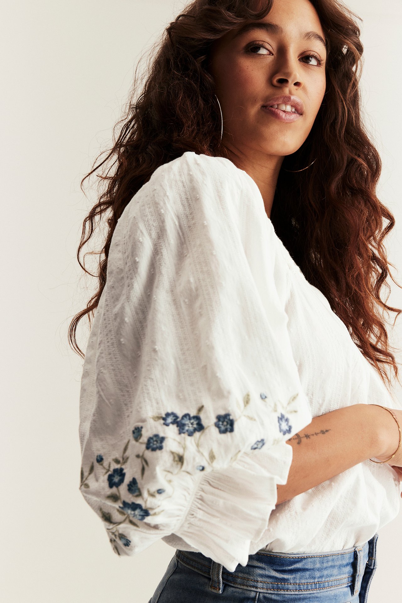 Nainen luonnonvalkoisessa puuvillapuserossa, jossa sinisiä kukkakirjontoja puhvihihoissa, ja sinisissä farkuissa.