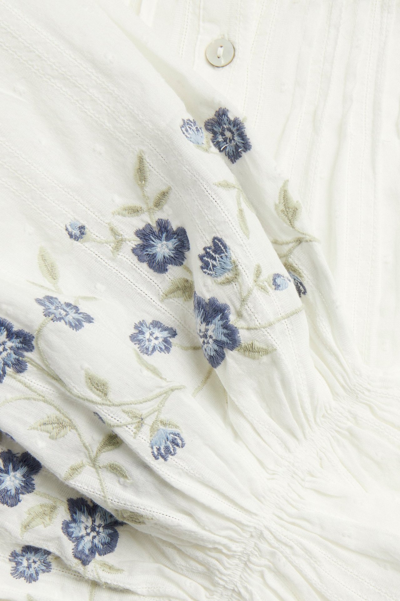 Nærbilde av en offwhite bomullsbluse for dame med blå blomsterbroderier, teksturerte striper og en synlig knapp.