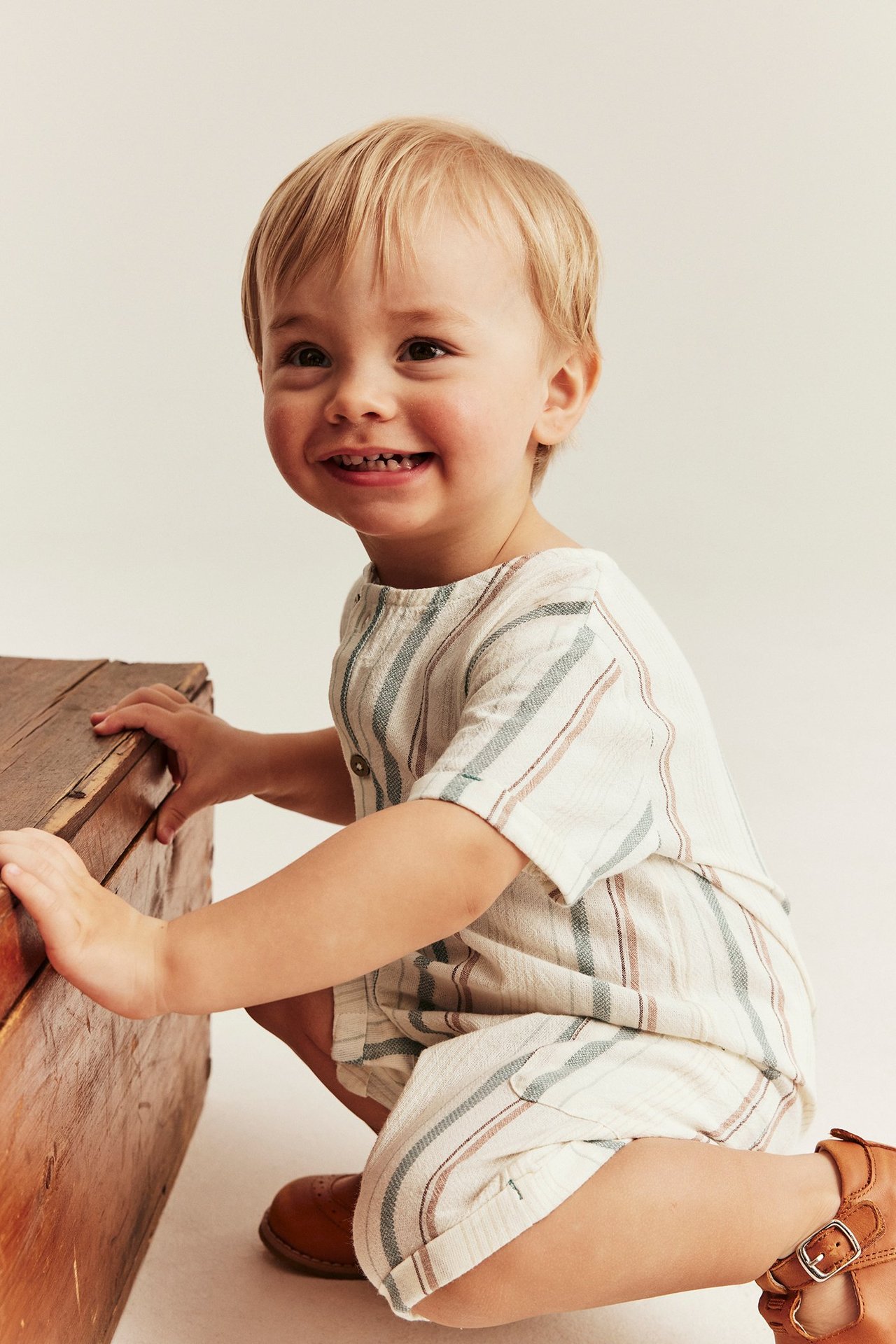 Smilende baby i offwhite stripet bomullsromper og brune sko, huker ved trekasse.