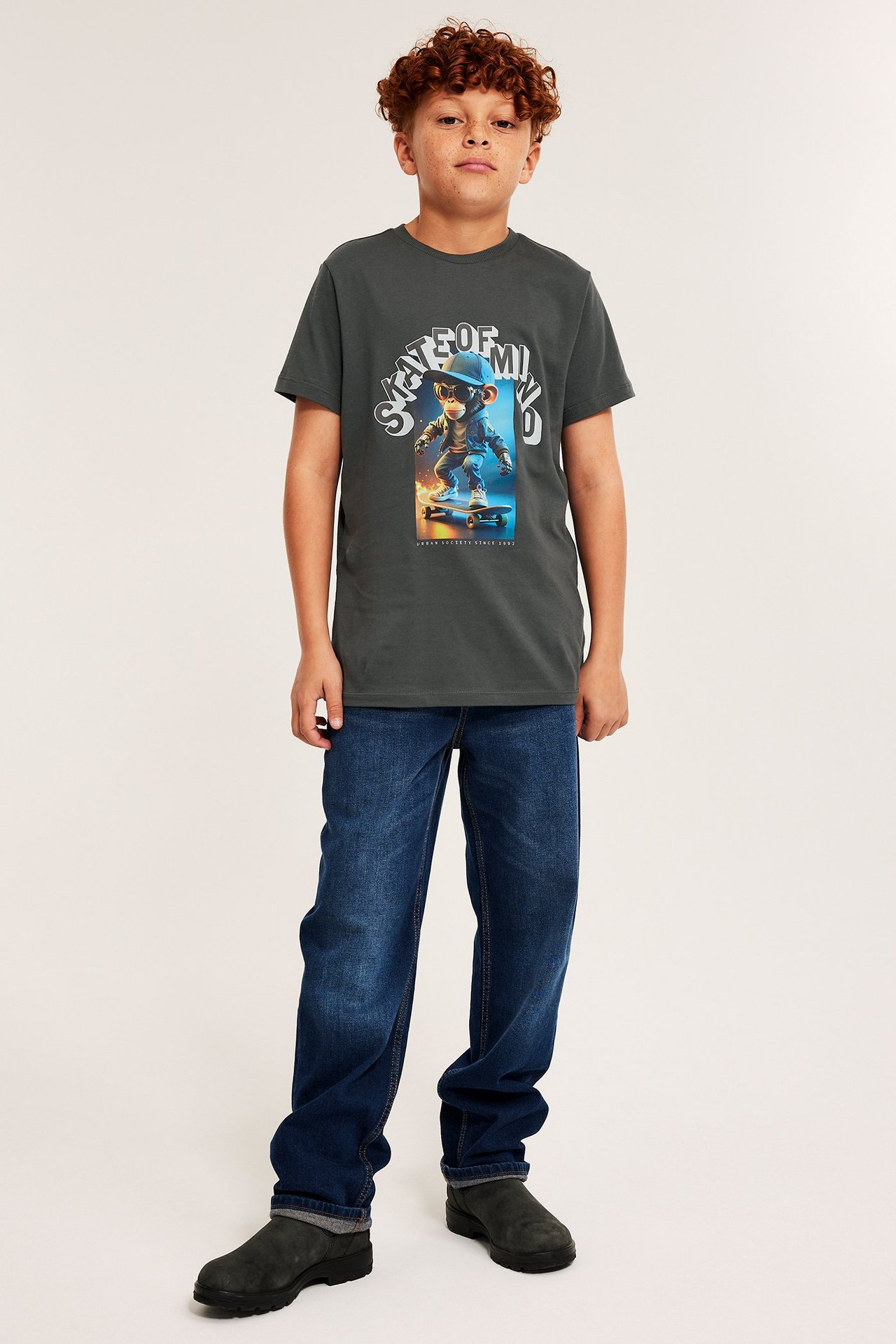 Barnkille i grå T-shirt med tryck fram, blå jeans och svarta skor, frontvy.