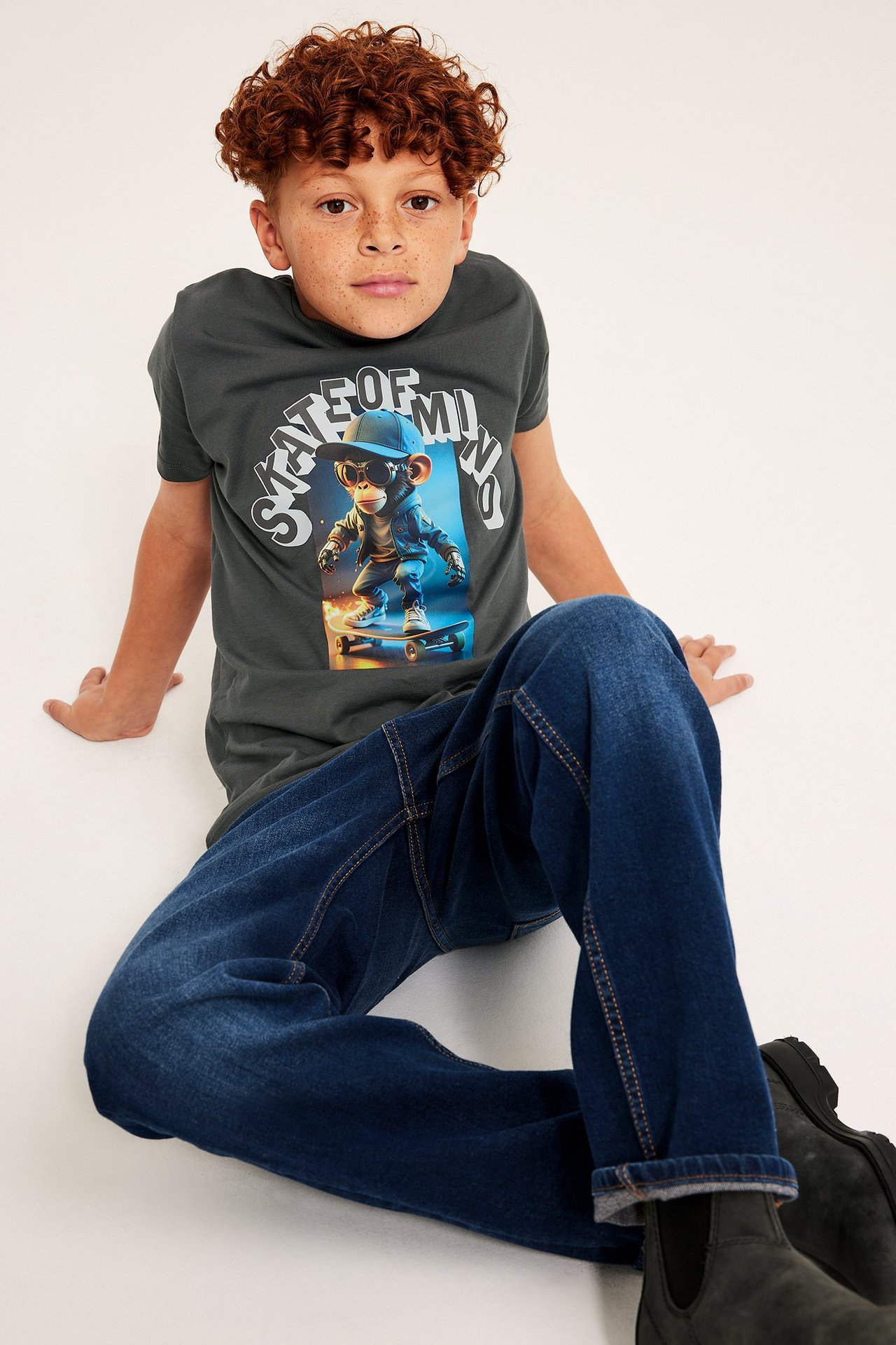 Ung man i grå t-shirt med tryck och blå jeans, sittande mot vit bakgrund.