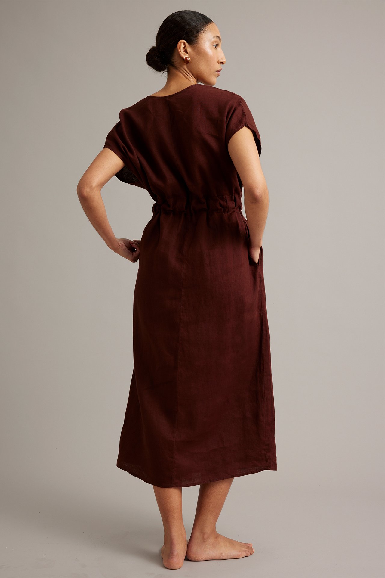 Sivulta takaa: Nainen yllä yksivärinen viininpunainen pellavamekko solmittavalla vyötäröllä ja sivutaskuilla.