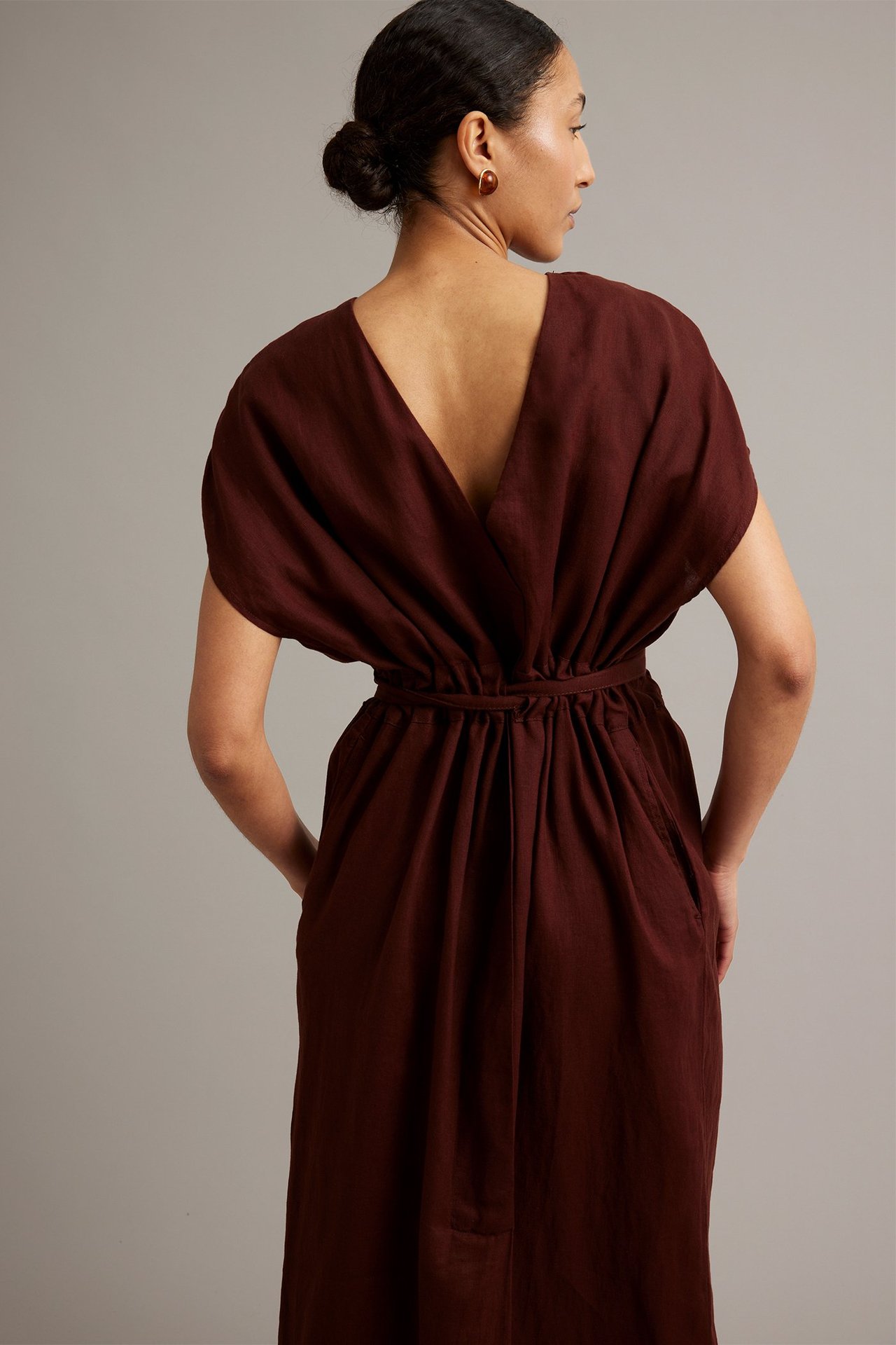 Takakuva: Nainen viininpunaisessa V-pääntie pellavamekossa, solmittu vyötärö, sivutaskut, ruskeat korvakorut.