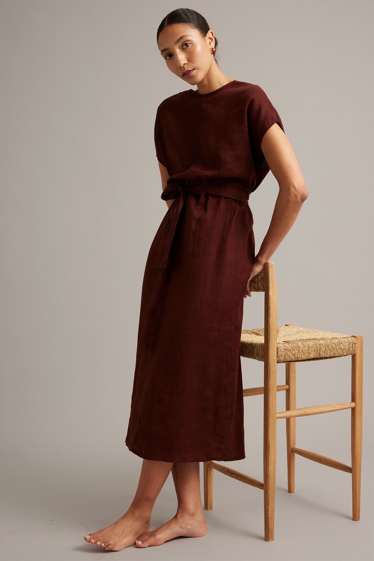 Edestä: Nainen viininpunaisessa pellavamidimekossa, jossa vyötäröllä sidottu vyö.