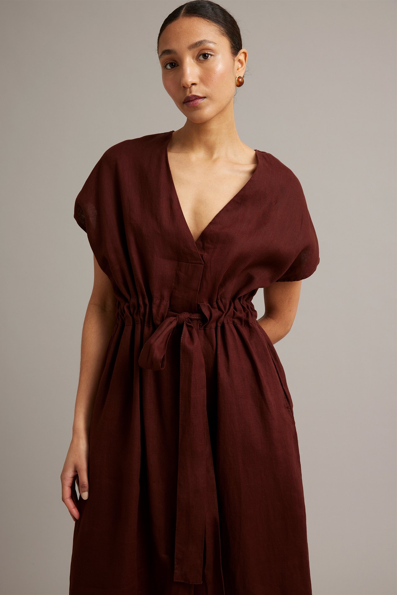 Framifrån, kvinna i vinröd linneklänning med V-ringning, knytband i midjan och sidfickor.