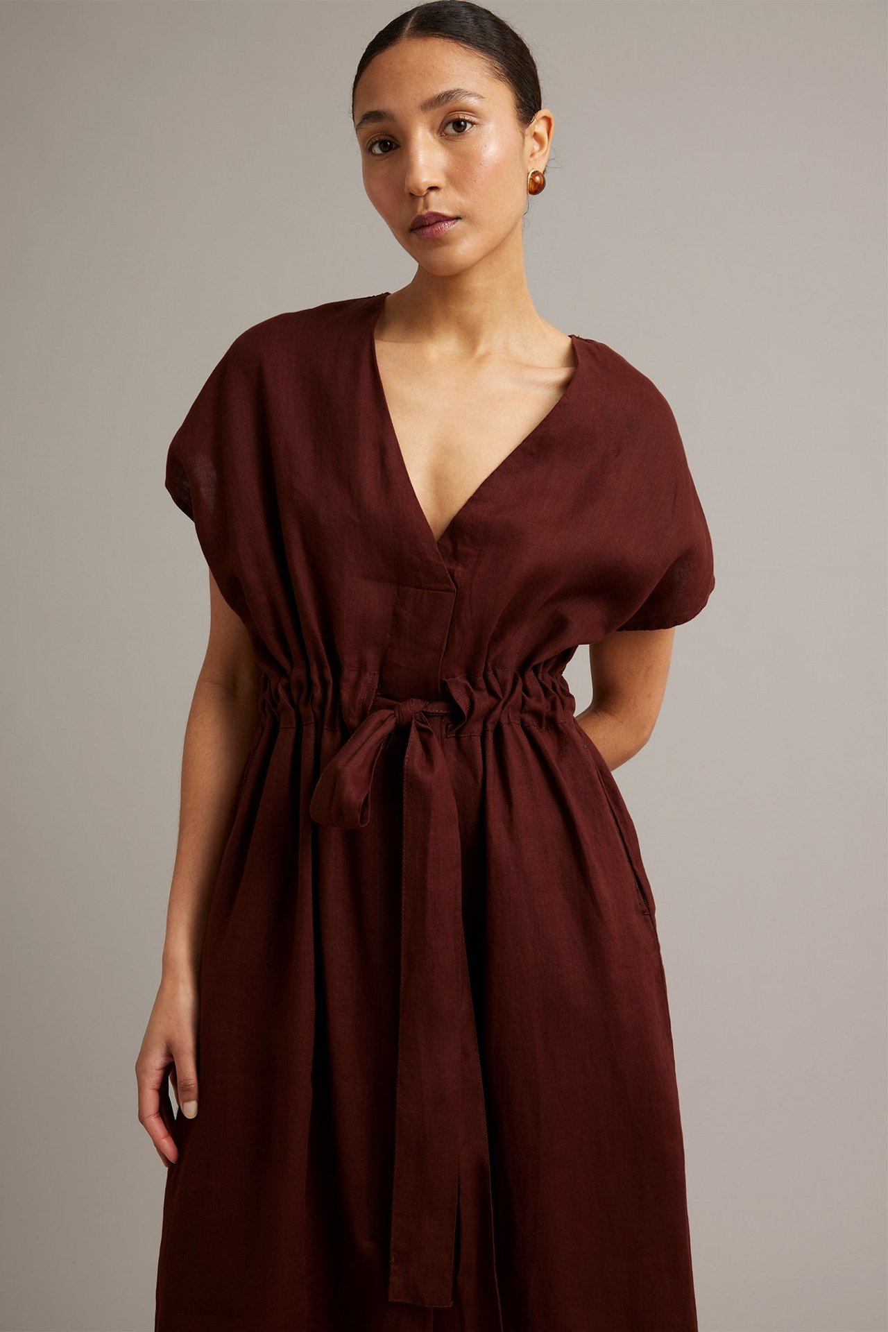 Widok z przodu, kobieta w bordowej lnianej sukience z dekoltem w serek, wiązanym paskiem i kieszeniami.