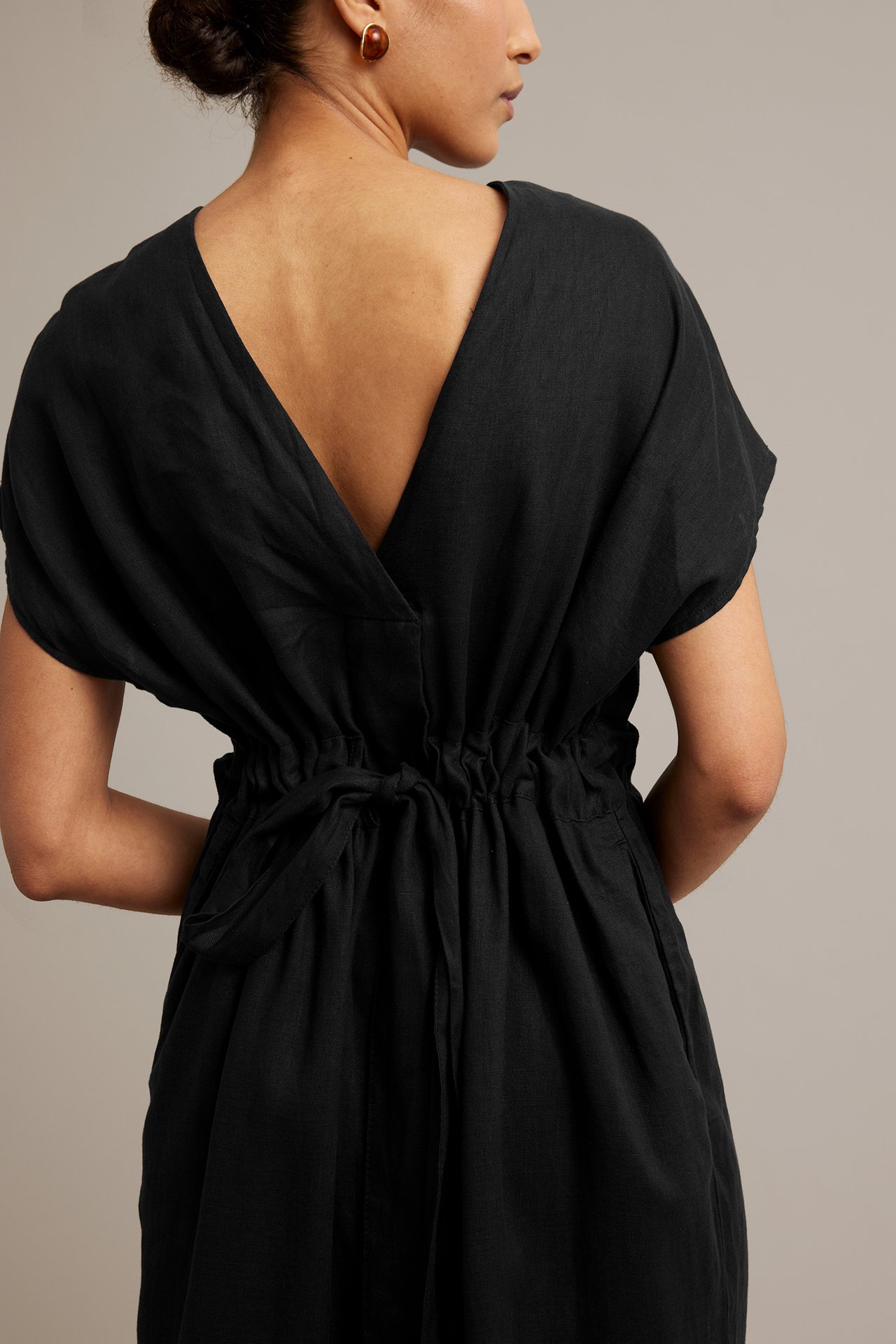 Widok z tyłu kobiety w czarnej lnianej sukience z dekoltem V, wiązaną talią i brązowymi kolczykami.