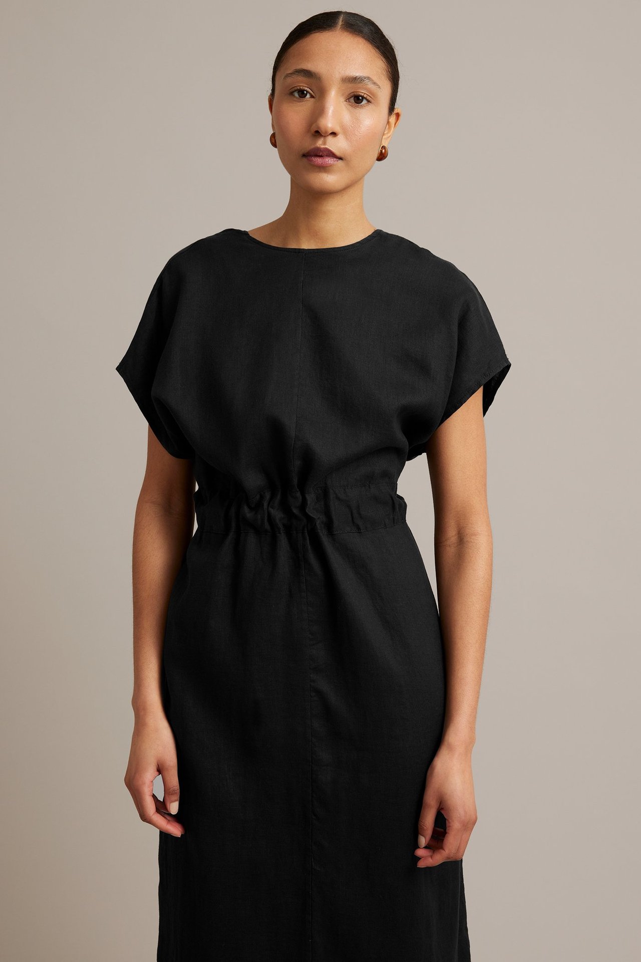 Widok z przodu: kobieta w czarnej lnianej sukience ze ściąganą talią, krótkimi rękawami i brązowymi kolczykami.