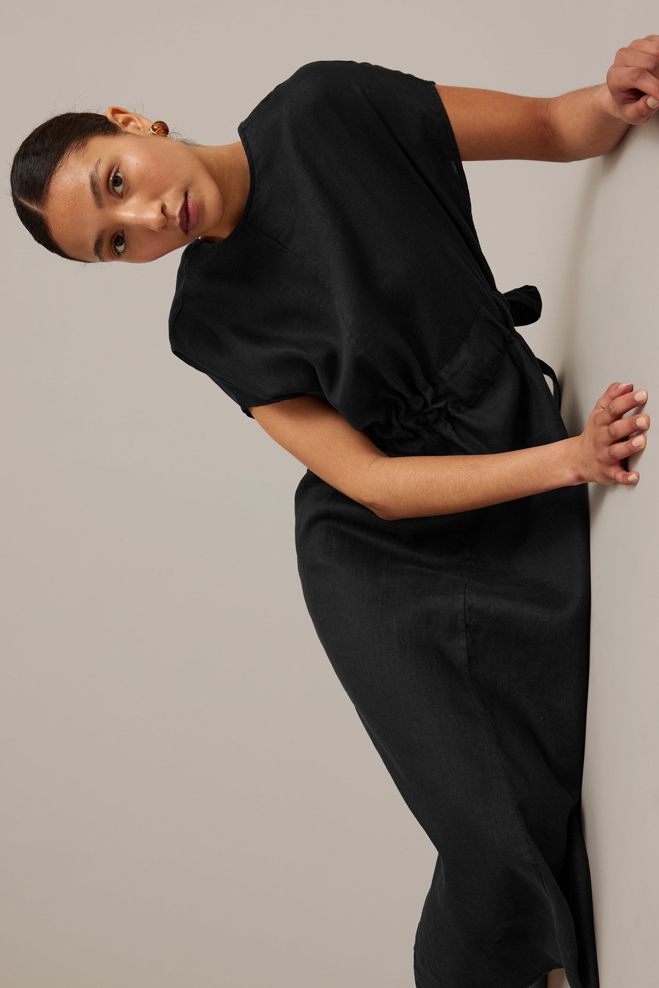 Frontal bild på en kvinna i en svart linneklänning med korta ärmar, knuten midja och små bruna örhängen.