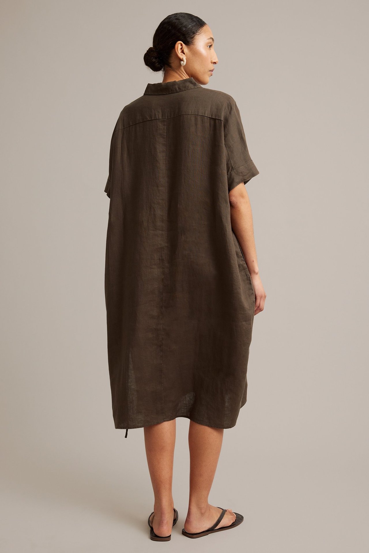 Bakfra, kvinne i en løs brun skjortekjole i lin med korte ermer og svarte flate sandaler.