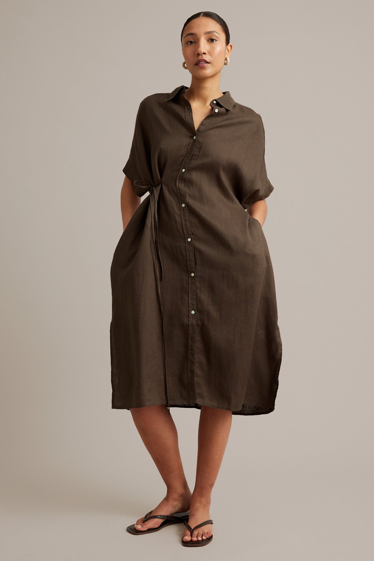 Front view: Kvinna i brun linneskjortklänning med knytband i midjan, sidofickor och svarta flip-flops.