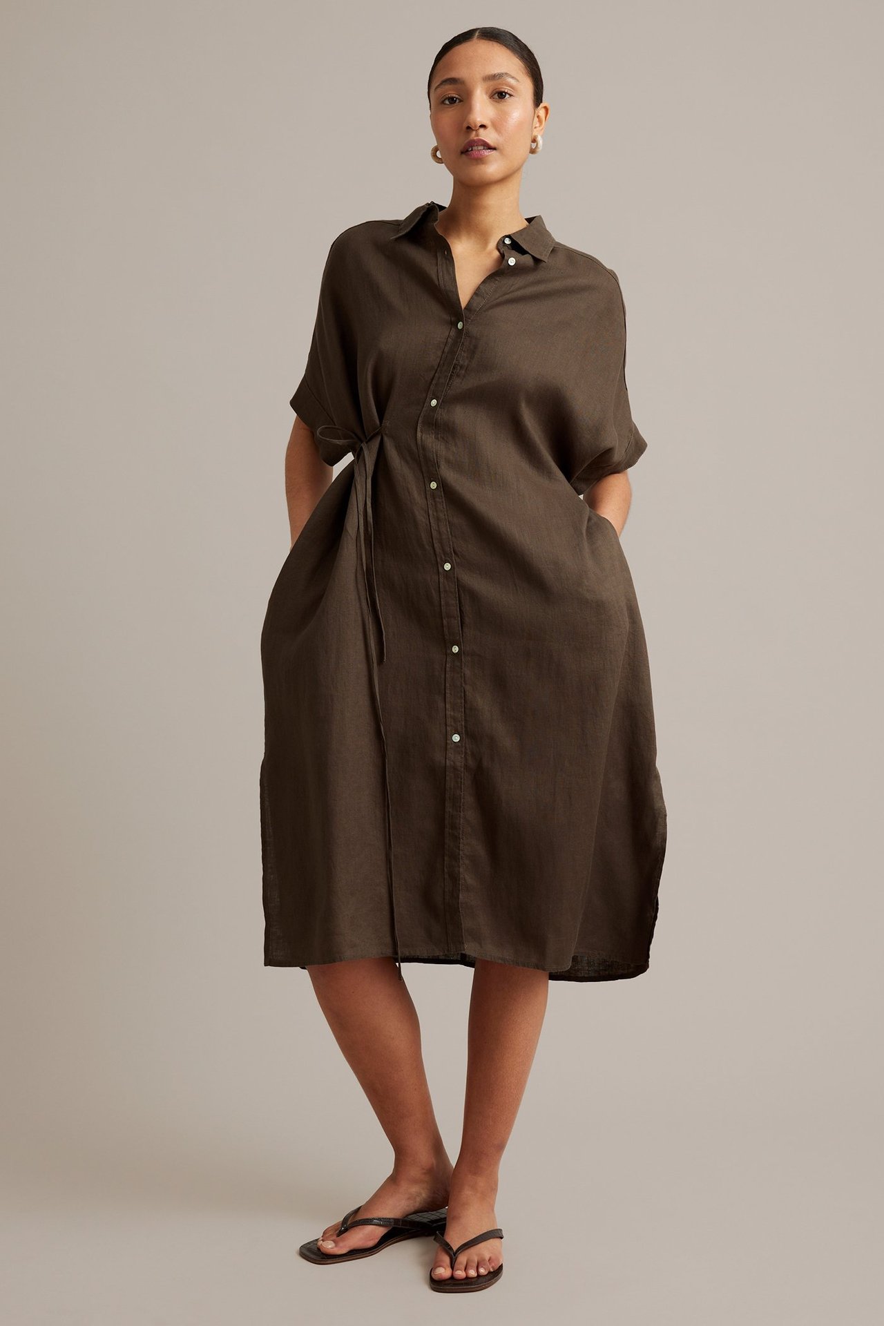 Front view: Kvinne i brun linneskjortekjole med knytebånd i midjen, sidelommer og svarte flip-flops.